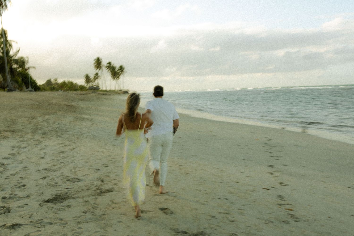 A couple in white attire walks together along a tropical beach at sunset with palm trees in the distance.