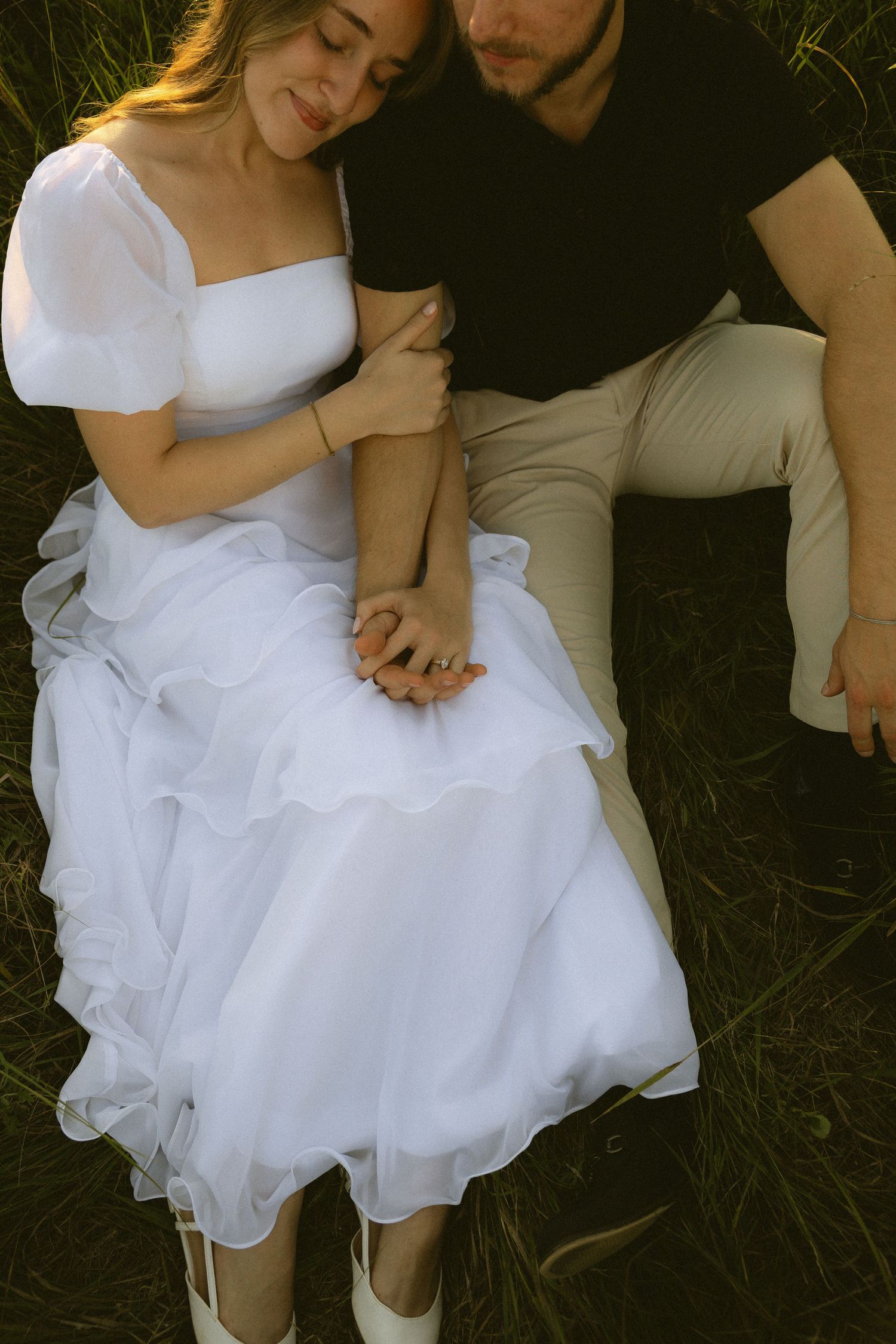 A bride in a white ruffled dress sits on grass holding hands with a man in a black shirt during an outdoor wedding.