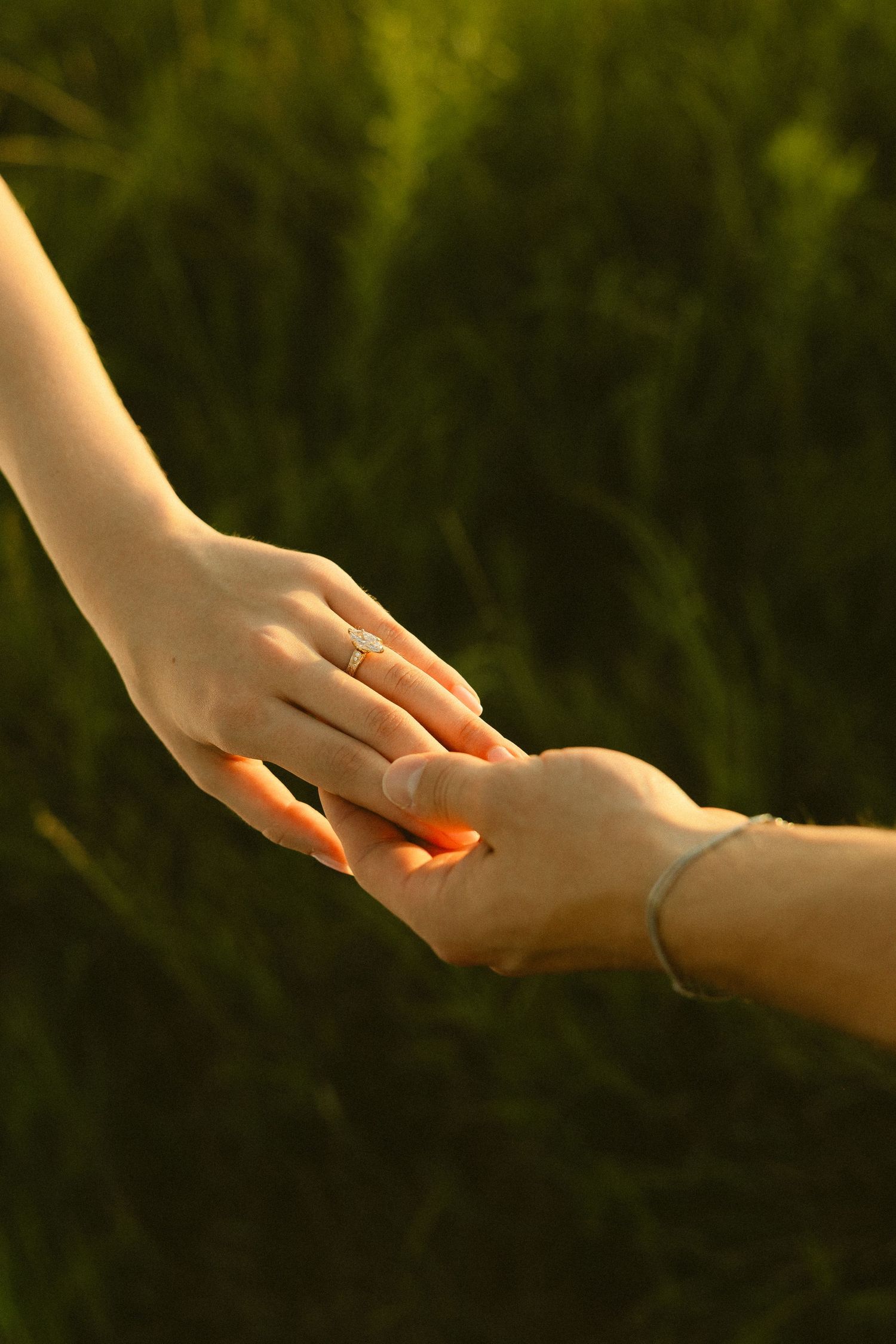Two hands gently touching at sunset with green grass background, woman wearing diamond engagement ring.