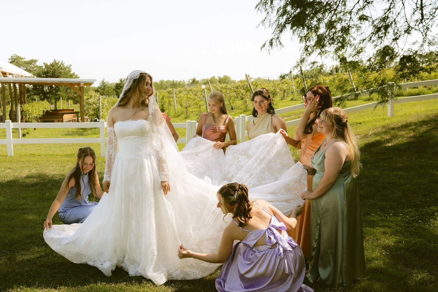 Bridesmaids helping bride arrange her white wedding gown train outdoors on sunny garden lawn.