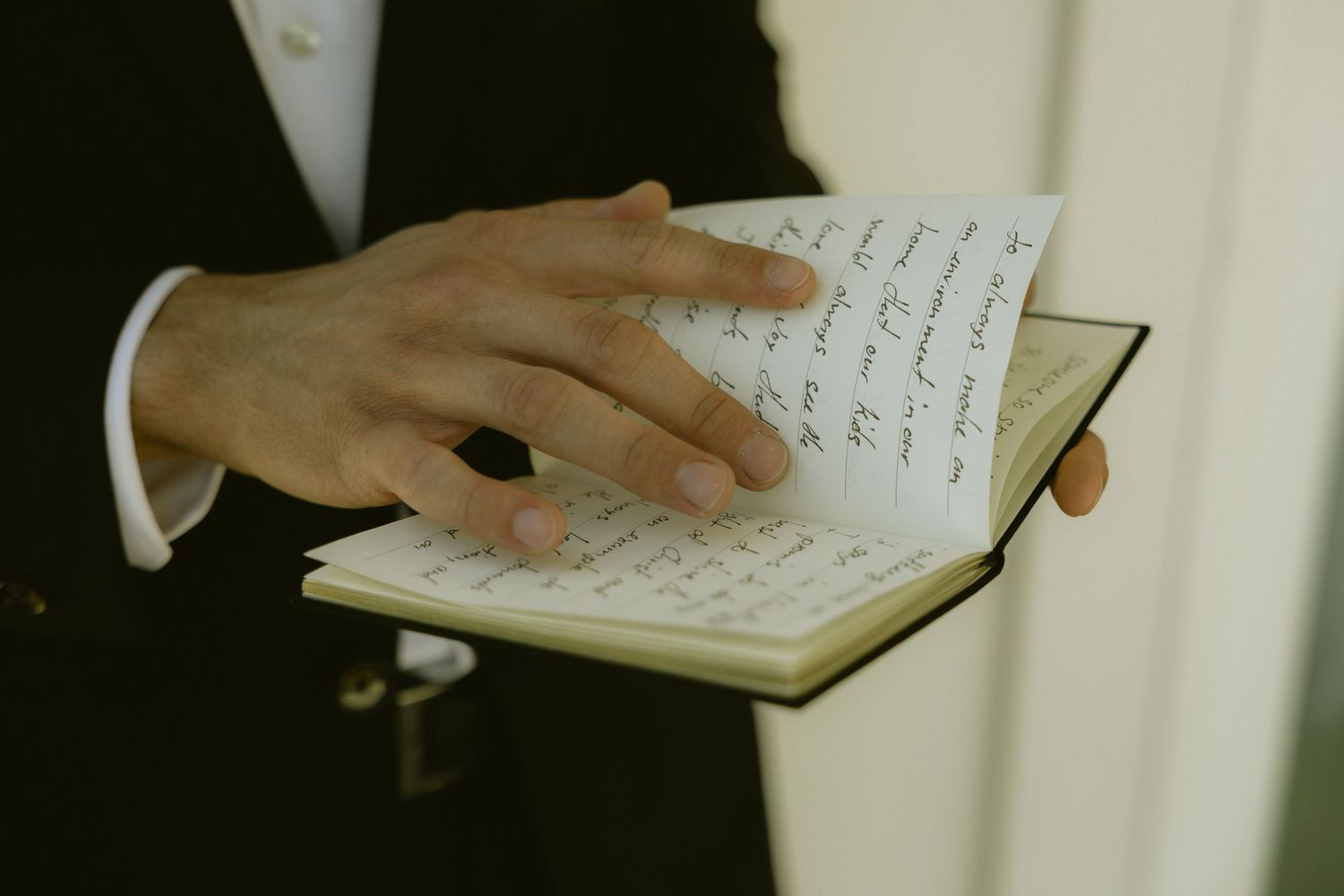 A groom in a tuxedo reads handwritten wedding vows from a small notebook during a ceremony.