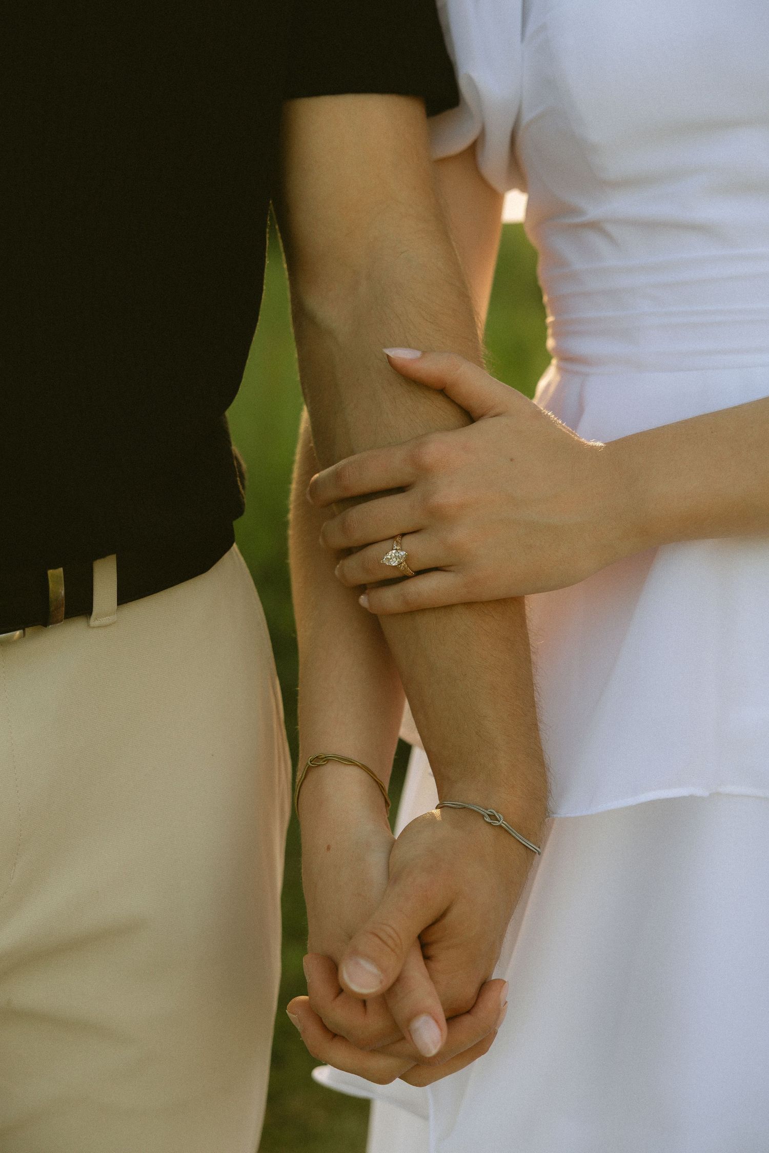 Couple holding hands, woman wearing diamond engagement ring and bracelet, man in khaki pants, outdoor golden hour setting.