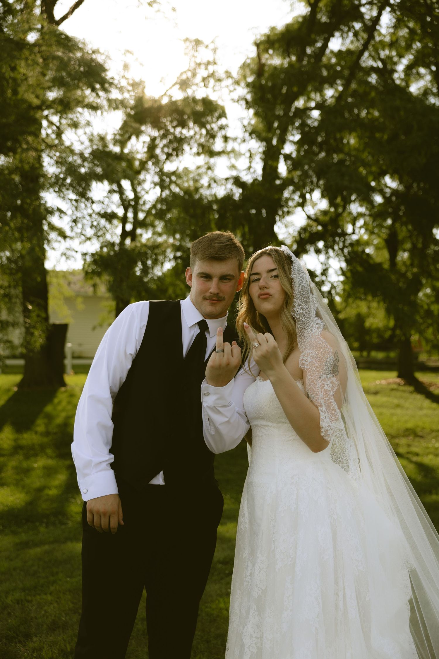 Bride and groom posing outdoors at golden hour, showing wedding rings, surrounded by lush green trees.