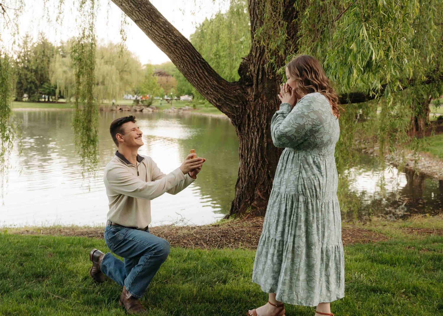 Man proposing on one knee to woman by a willow tree near a pond in a lush green park setting.