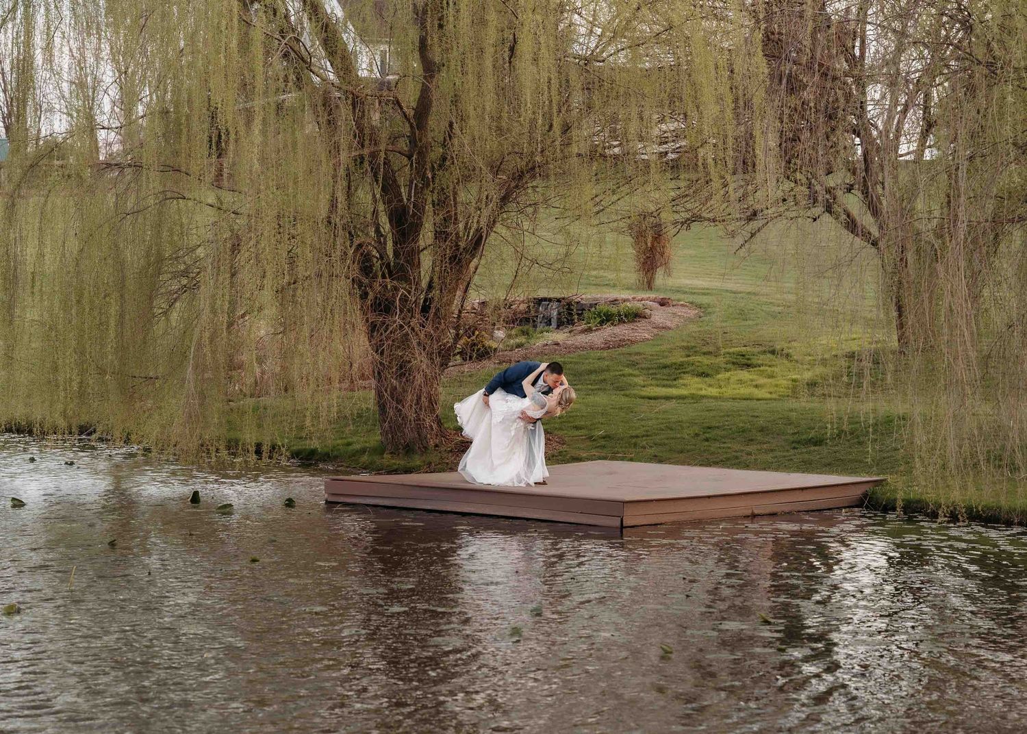 Couple sharing a romantic dip kiss on a floating dock surrounded by weeping willows and a serene pond.