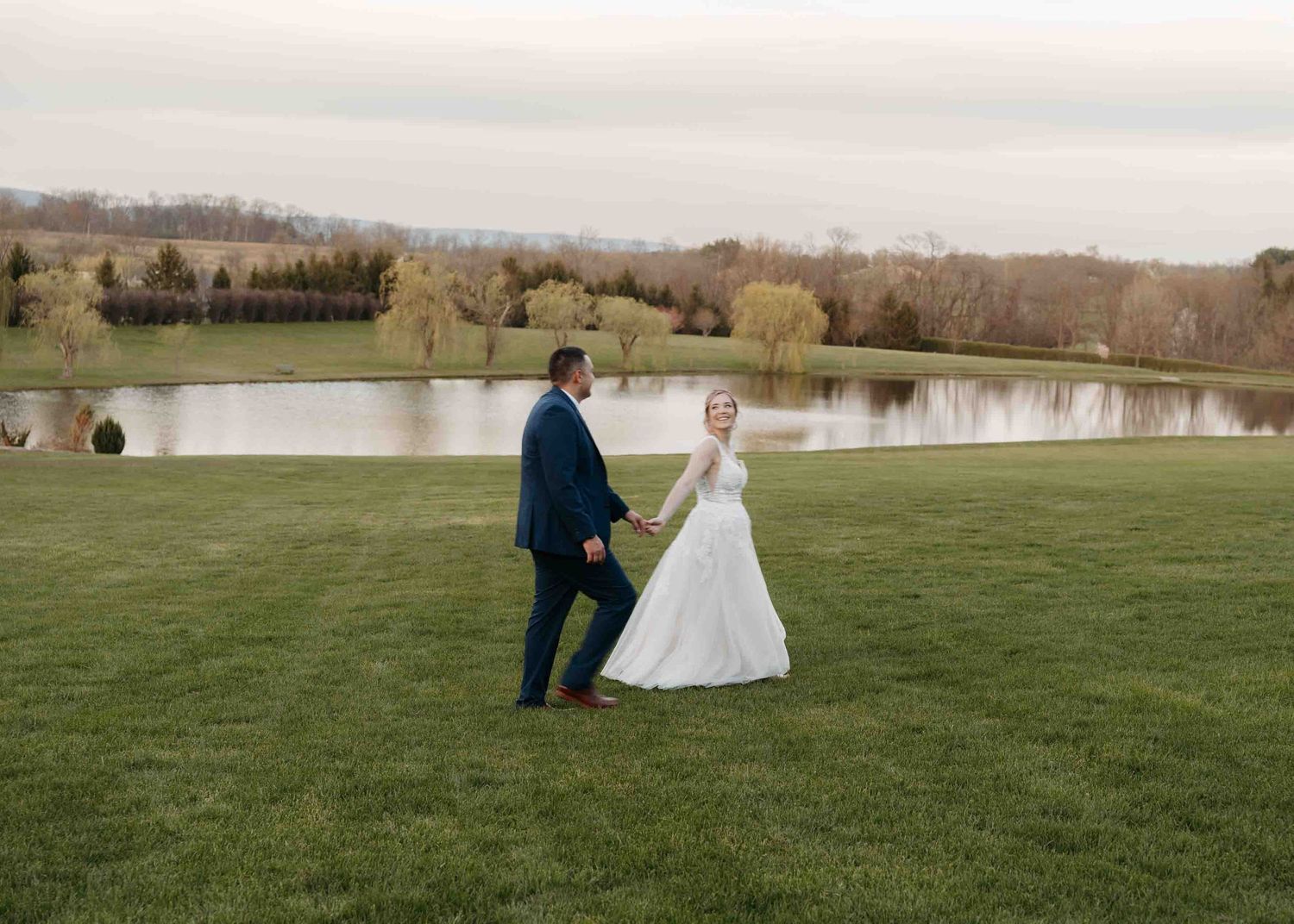 A bride and groom hold hands walking across lush green grass beside a serene pond at an outdoor wedding venue.