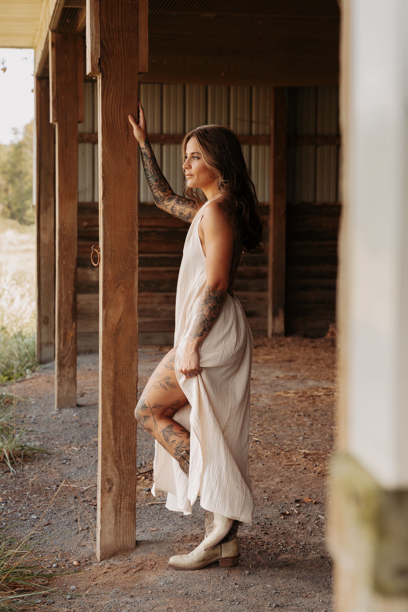 A figure in a flowing white dress poses gracefully against rustic wooden pillars in a dreamy outdoor setting.