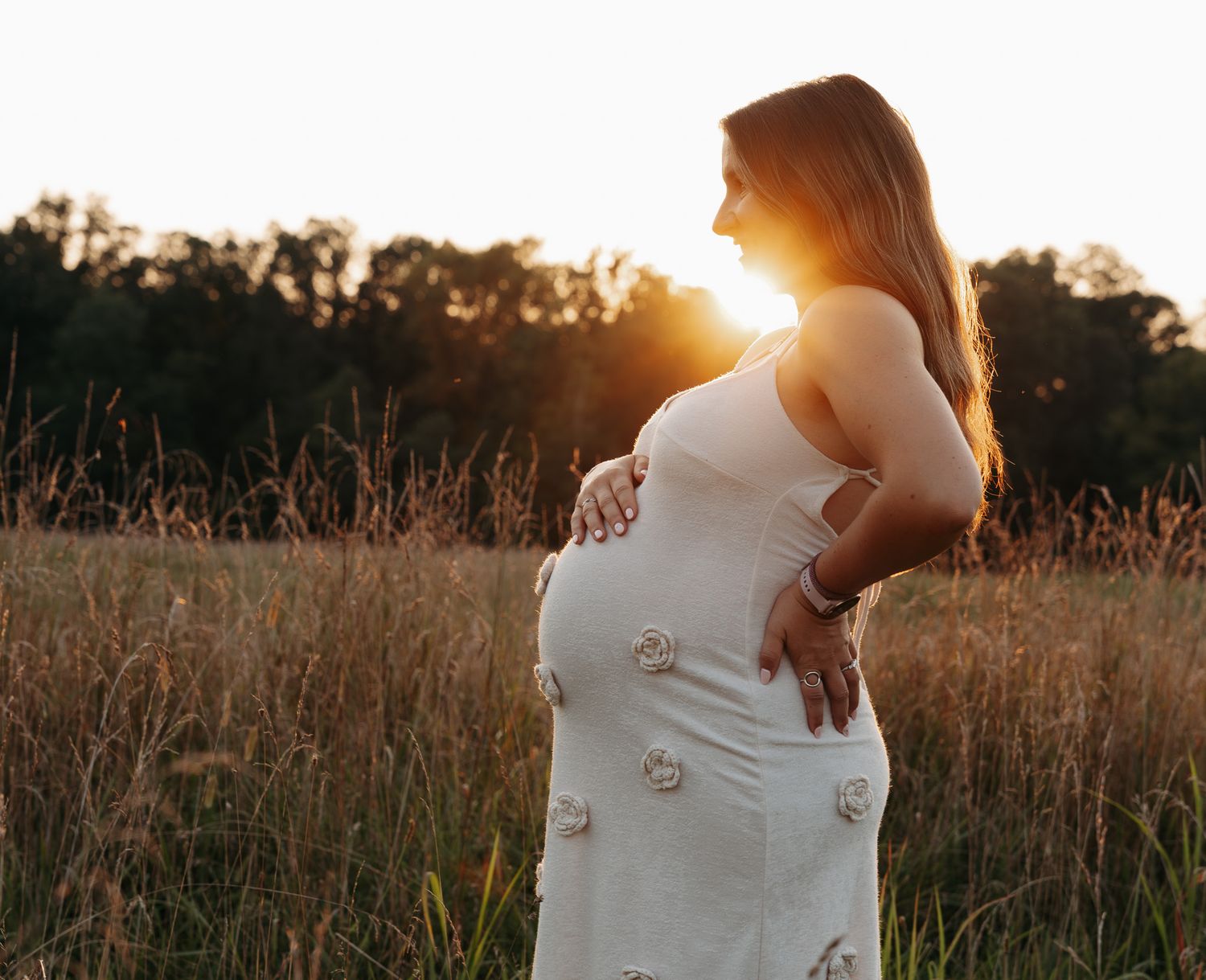 A pregnant woman in a white dress stands in a golden field at sunset with sunlight creating a warm silhouette.