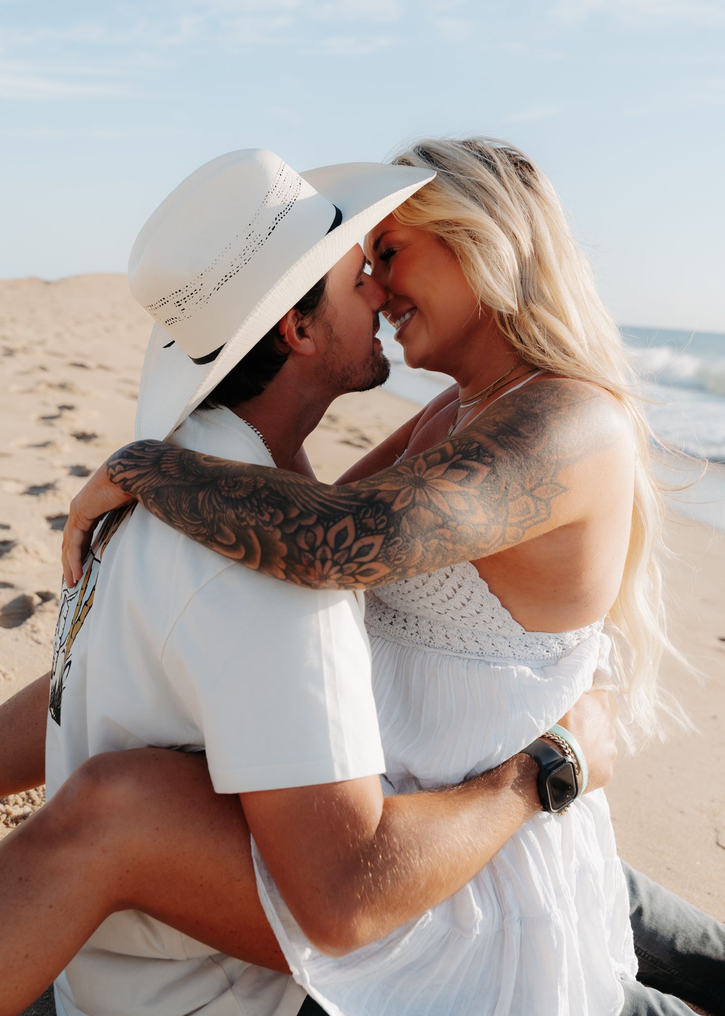 A romantic couple in white outfits share an intimate moment on a sunny beach, with visible tattoos and a summer hat.