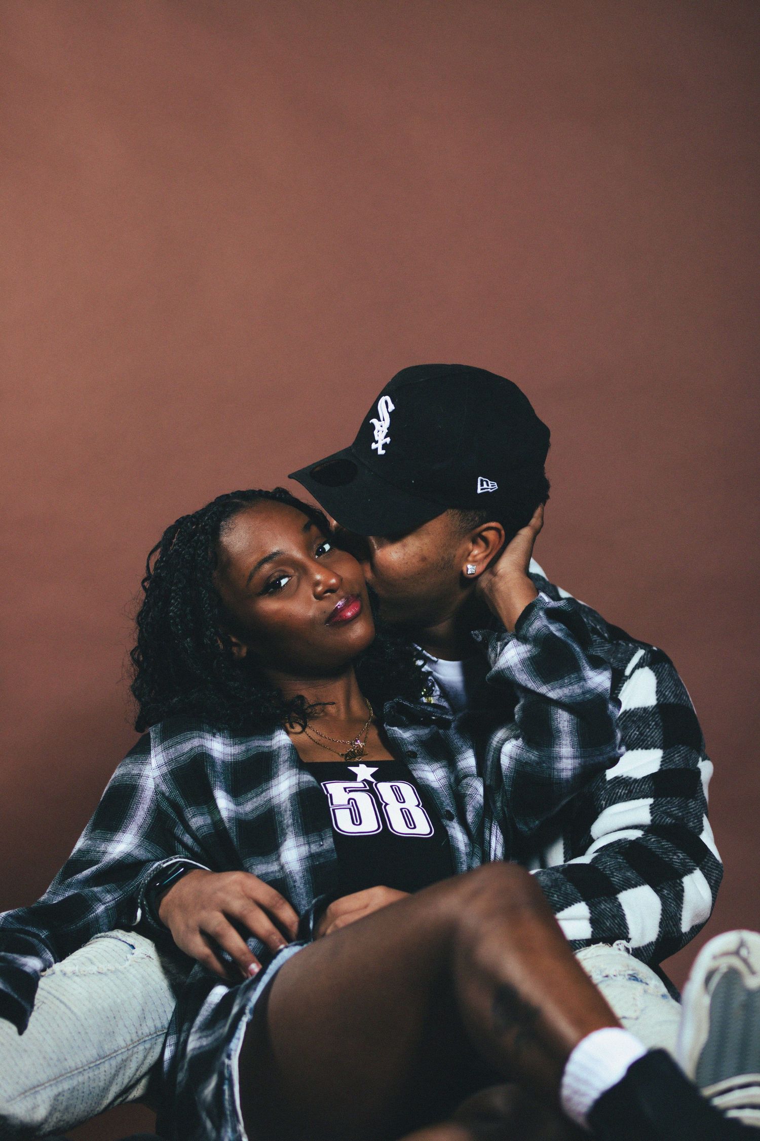 Couple in matching plaid shirts sitting together, man kissing woman on cheek against brown background.