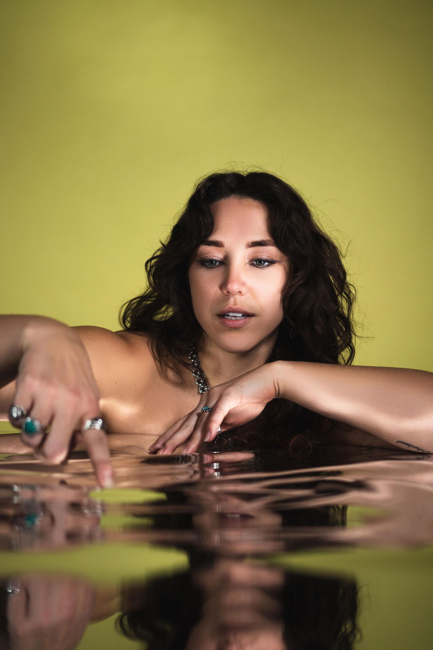 Woman with dark curly hair and turquoise ring rests arms on reflective water surface.