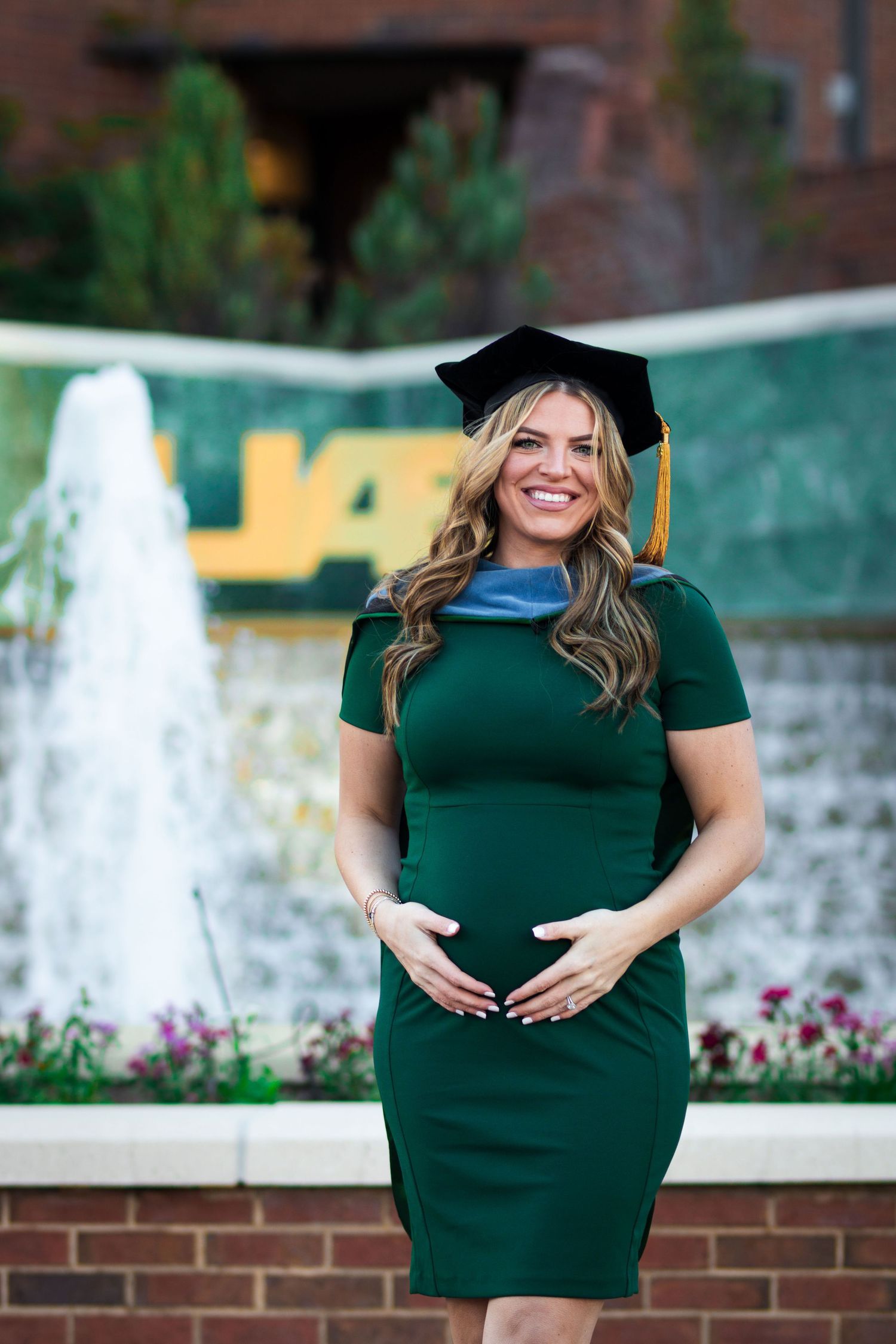 Pregnant female graduate in green dress and cap poses by UAB fountain.