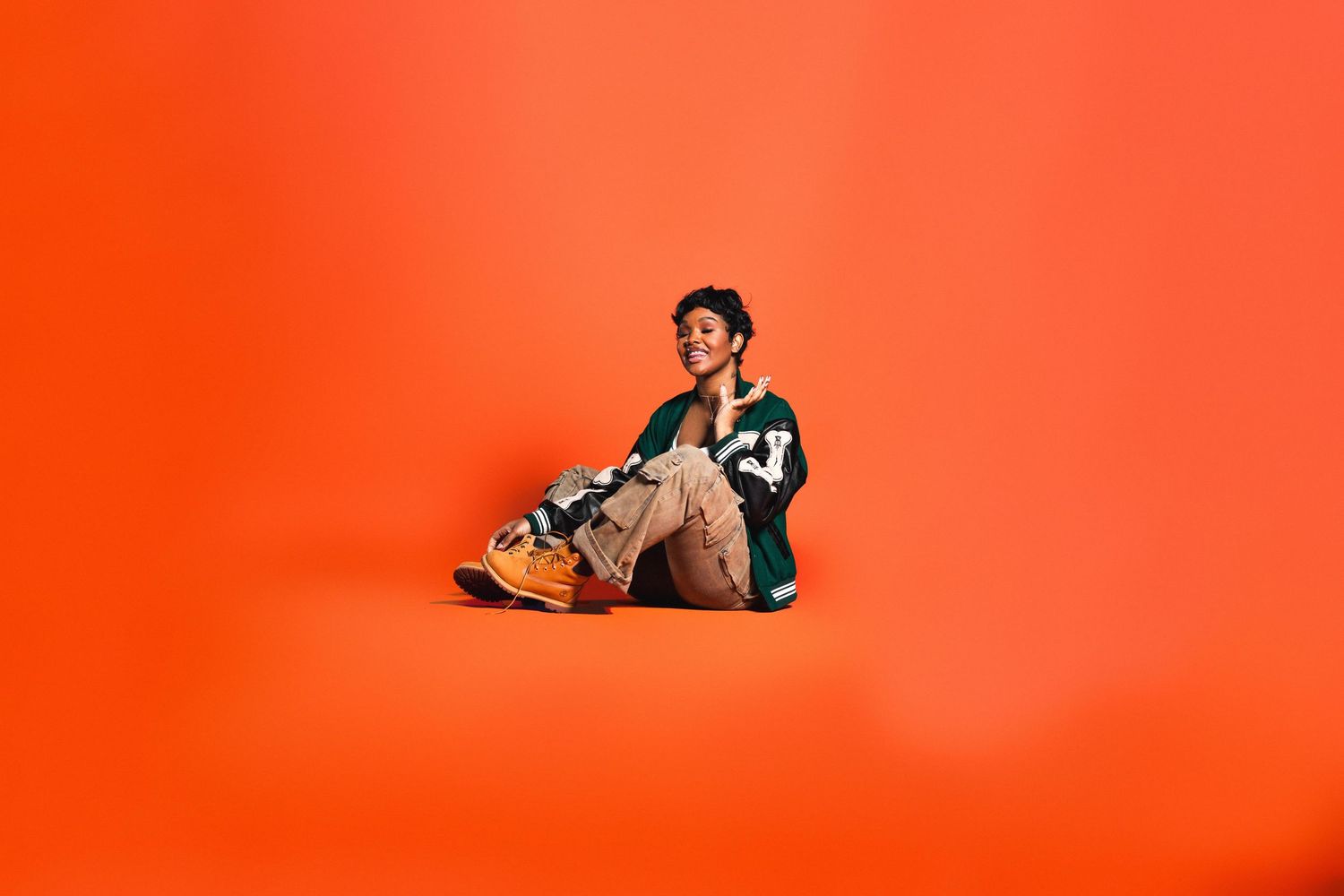 Young person in green varsity jacket and cargo pants sitting on floor against orange background.