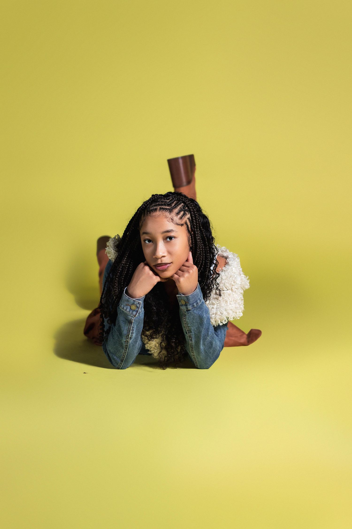 A young Black woman with box braids lies on the floor posing casually on a yellow-green background.