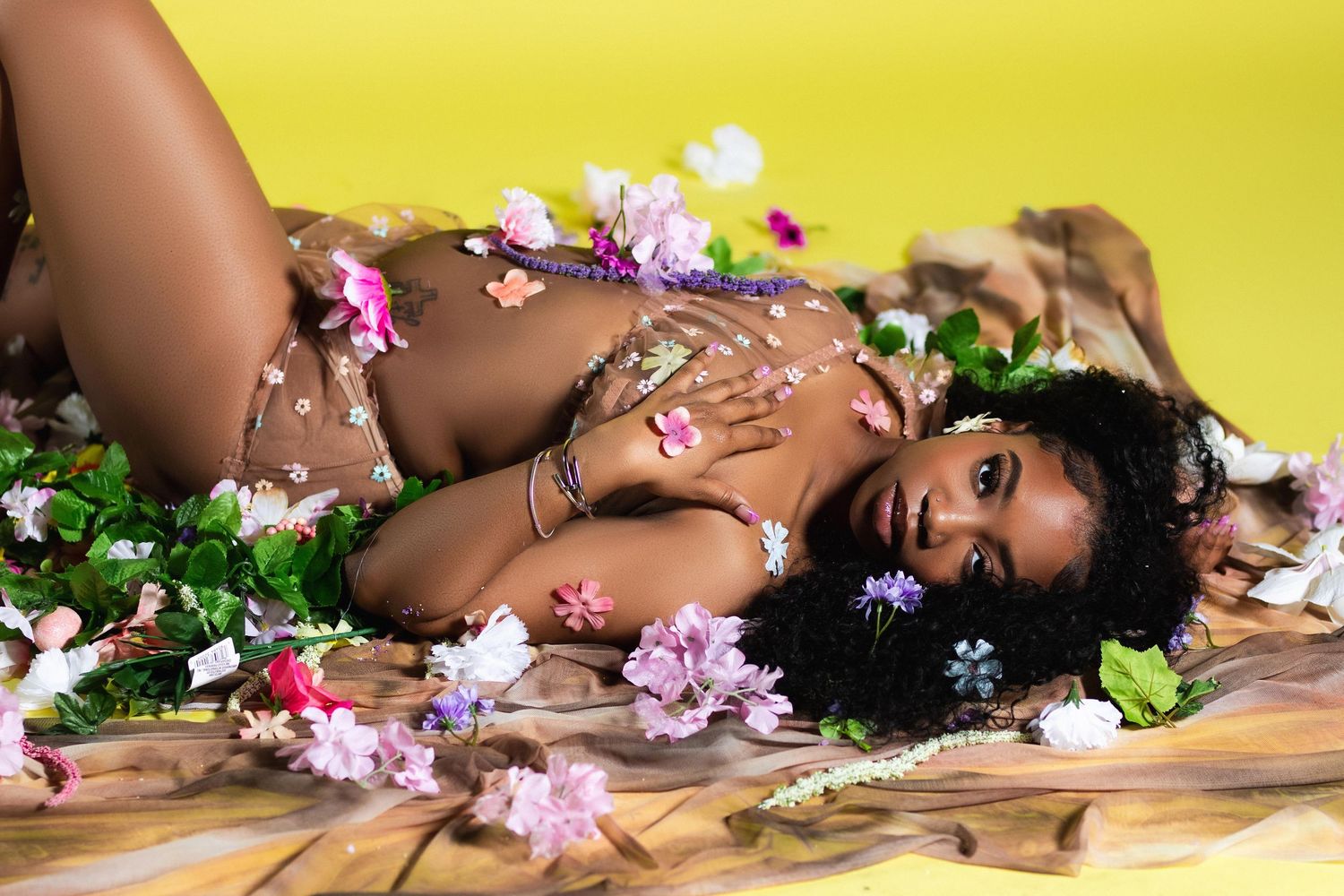 Woman lying among pink and purple flowers wearing floral accessories against a yellow background.