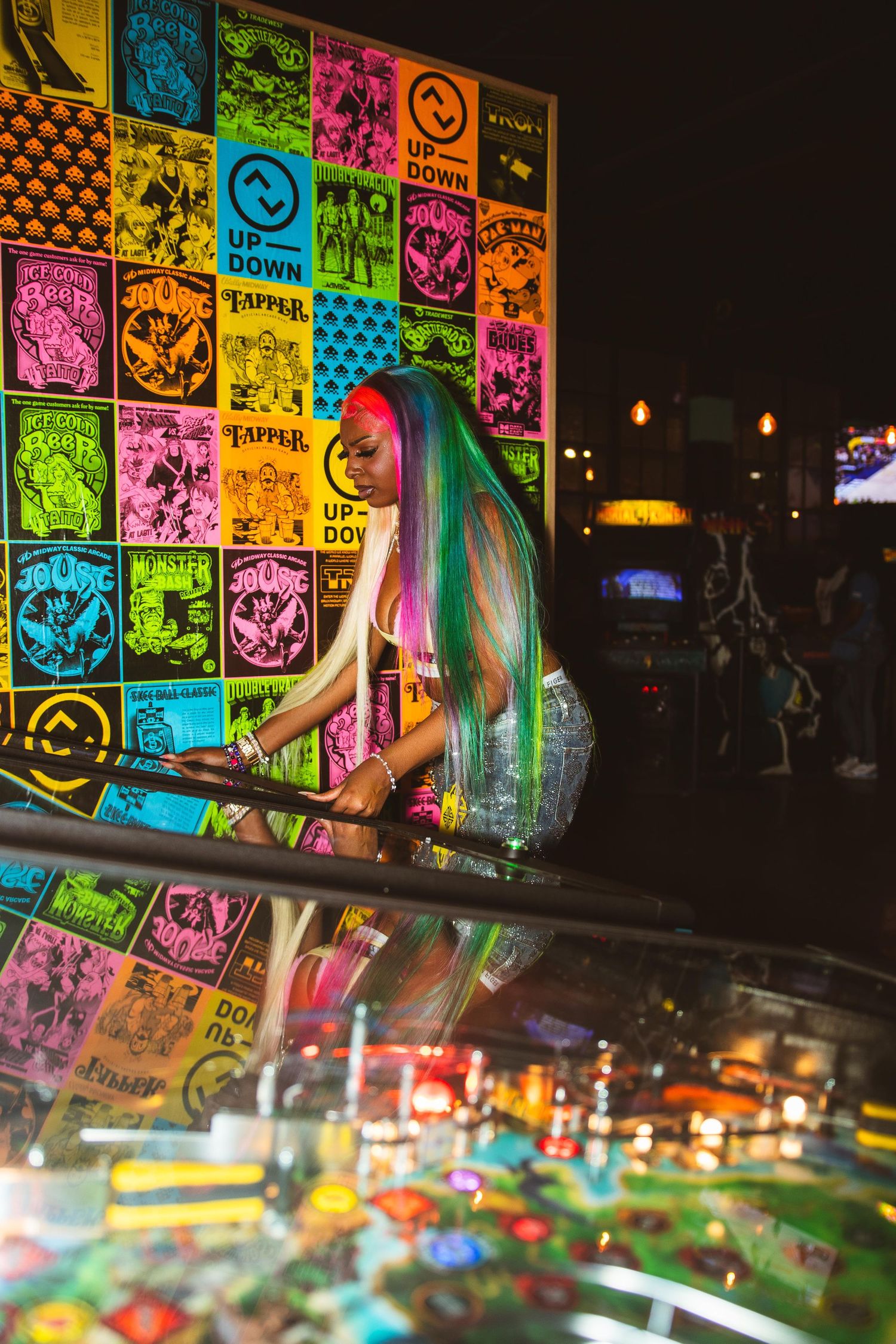 Woman with colorful hair plays pinball machine in front of vibrant neon arcade poster wall.