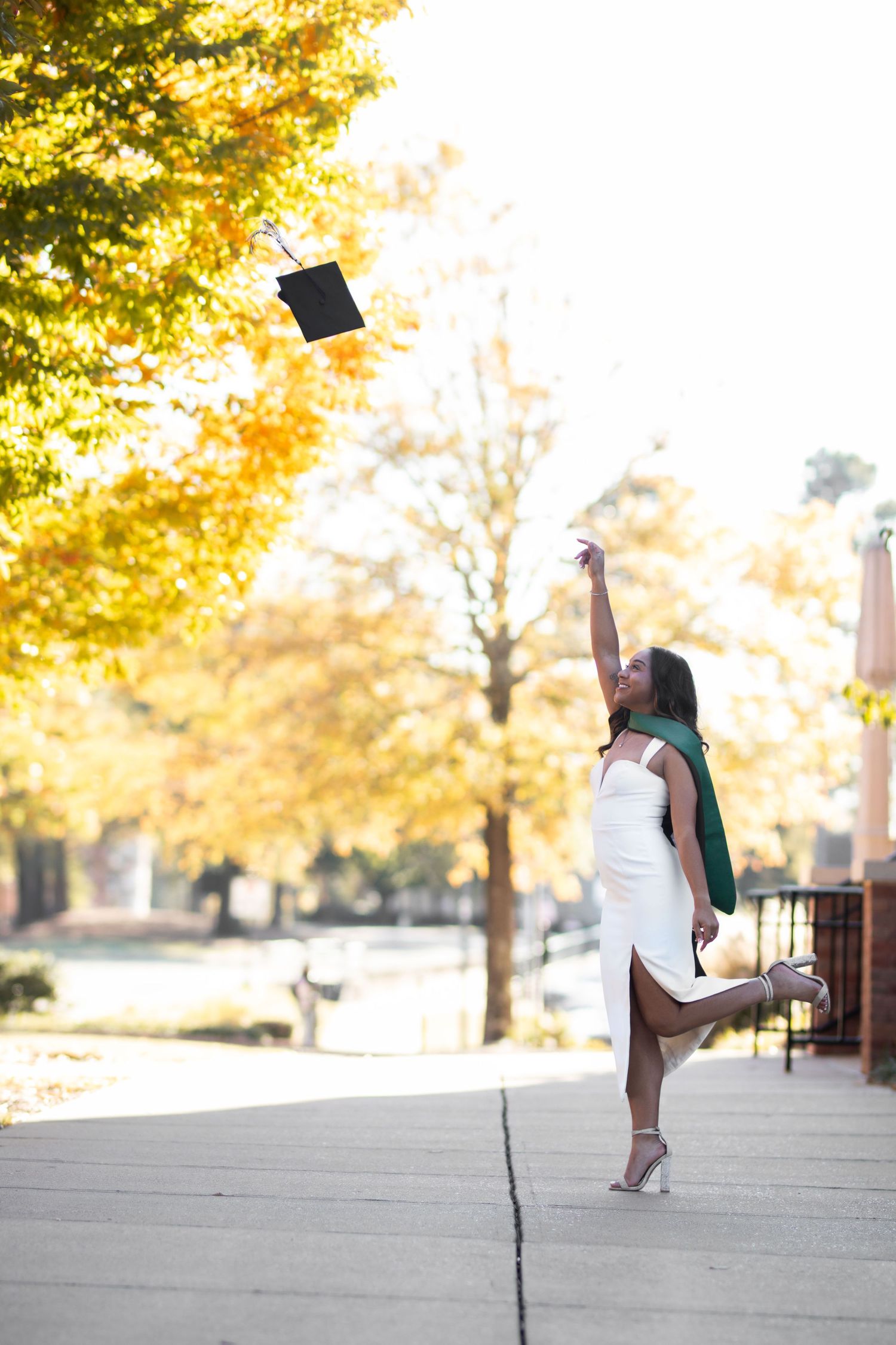 Young Black woman in white dress tossing black graduation cap into autumn foliage-lined walkway.