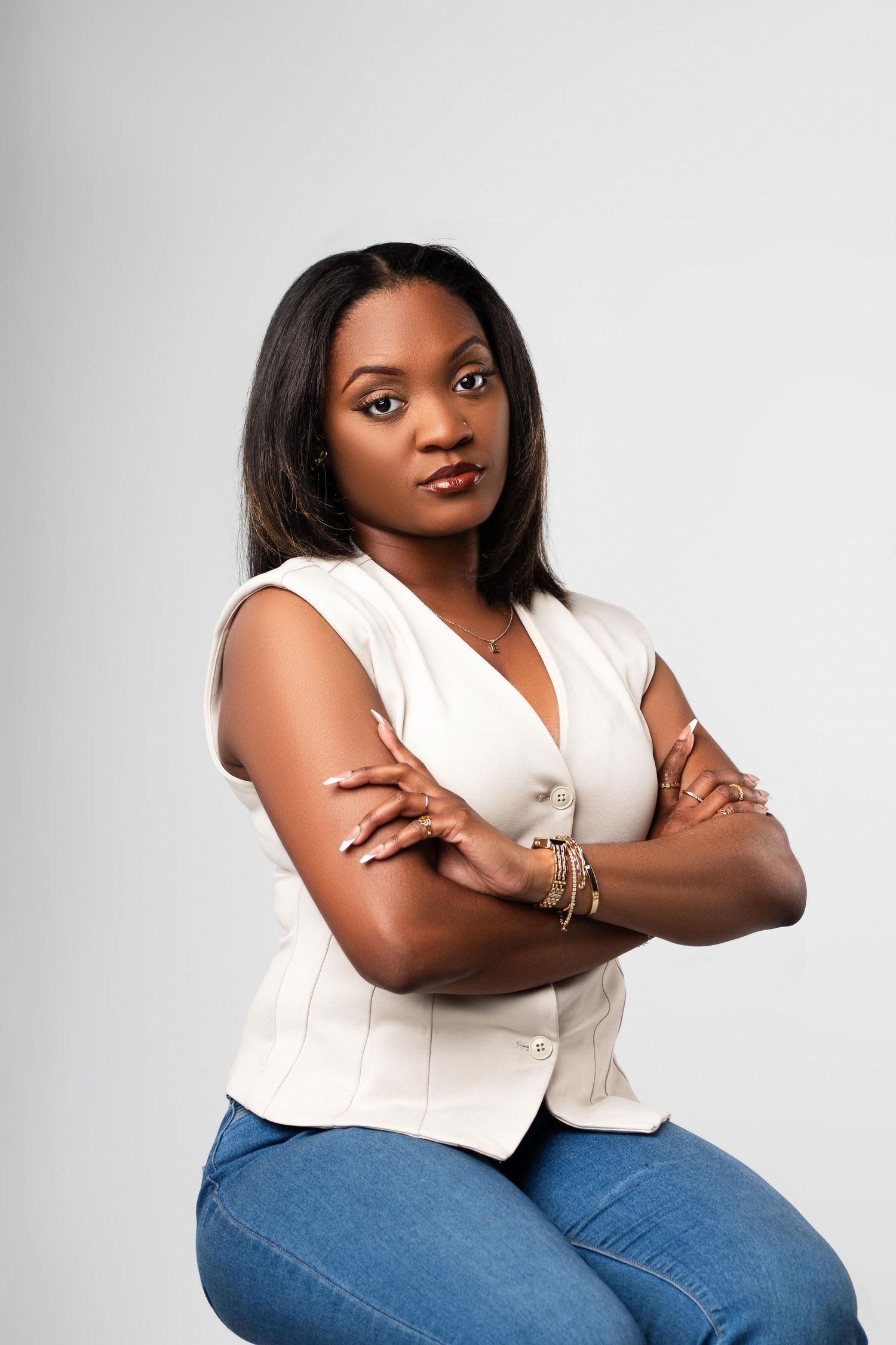 A confident Black woman in a white sleeveless top and jeans sits with arms crossed, posing against a gray background.