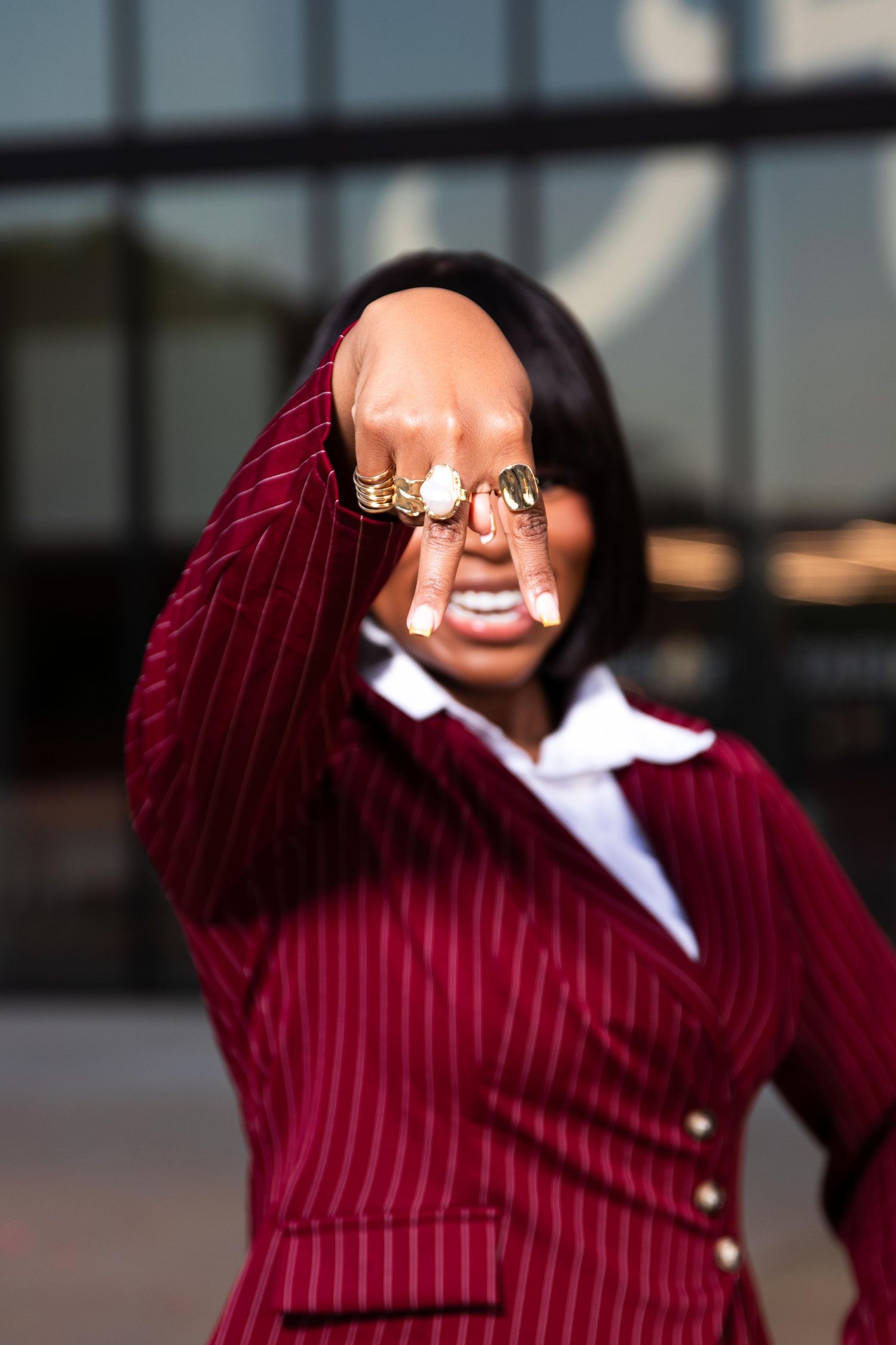 Woman in red pinstripe blazer confidently extends fist adorned with gold rings toward camera outdoors.