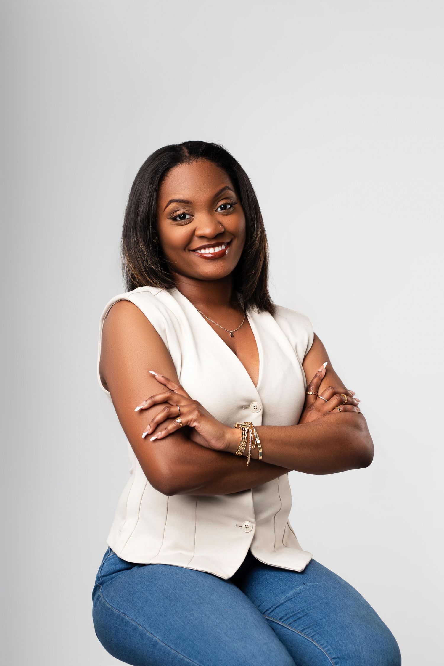 Smiling Black woman in white sleeveless top and jeans, arms crossed, posing confidently against a gray background.