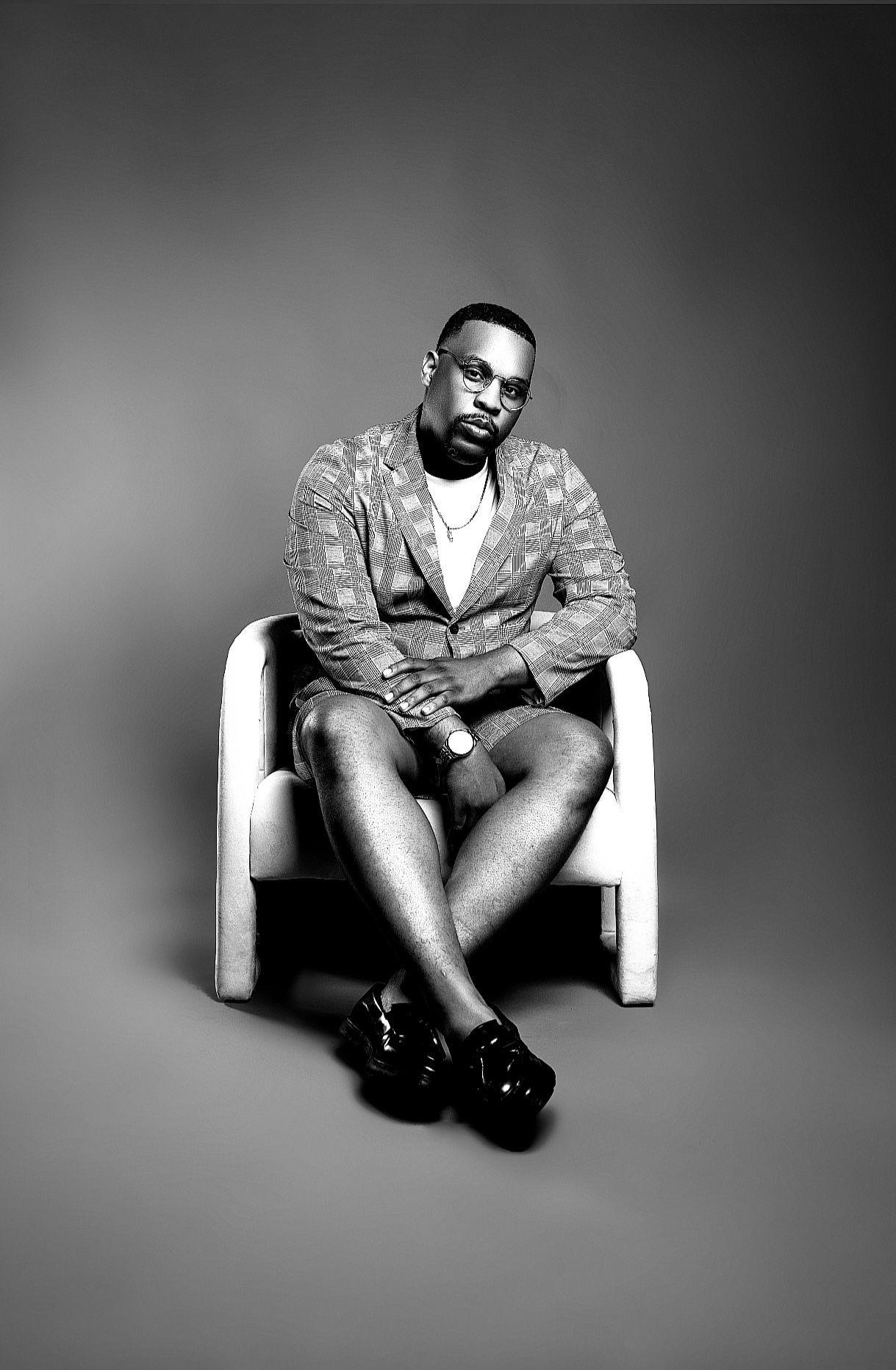 Stylish man in patterned blazer sits cross-legged in white chair in black and white portrait.