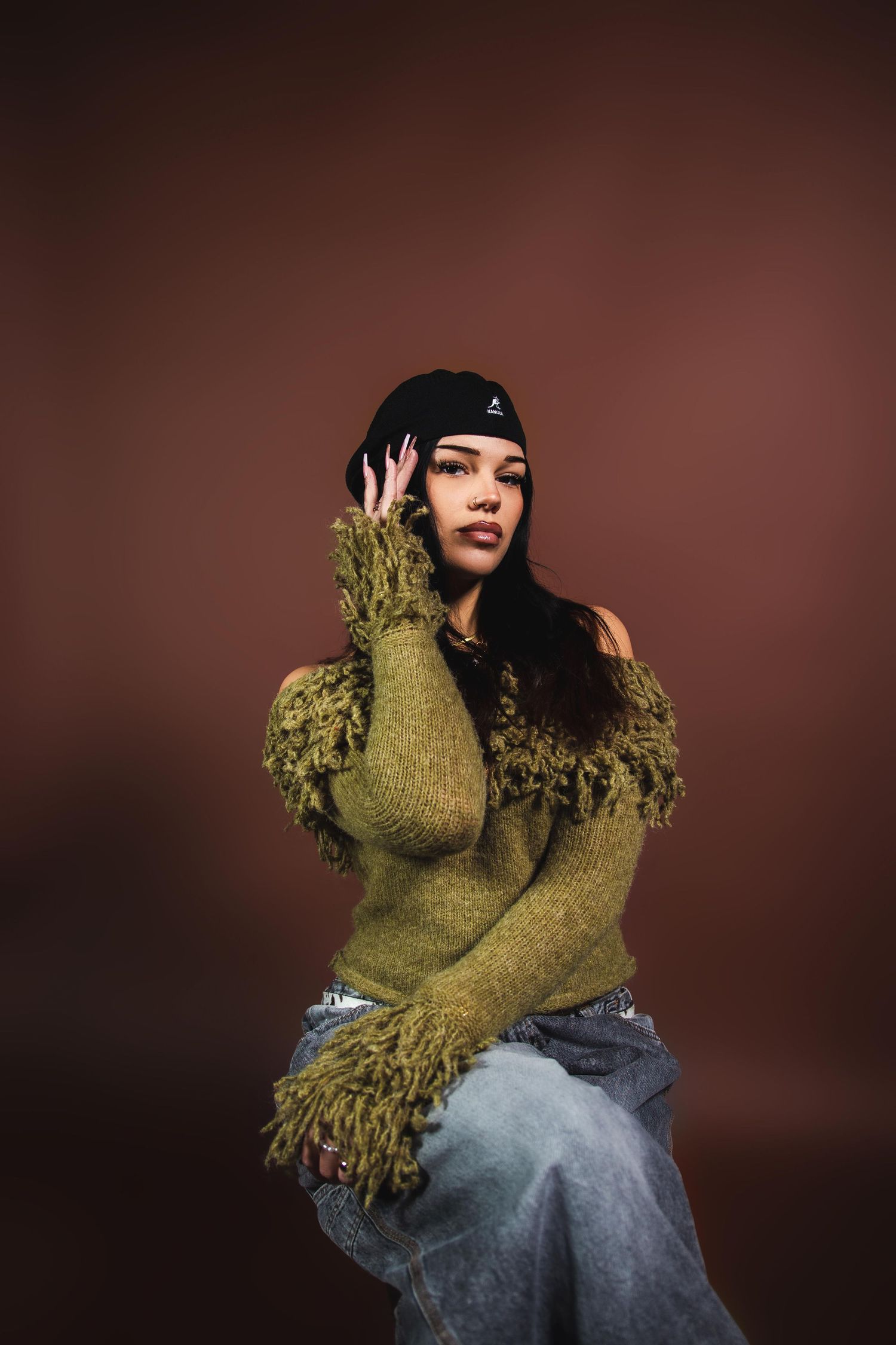 Woman in olive fringe sweater and grey jeans wearing black beanie, sitting against brown background.