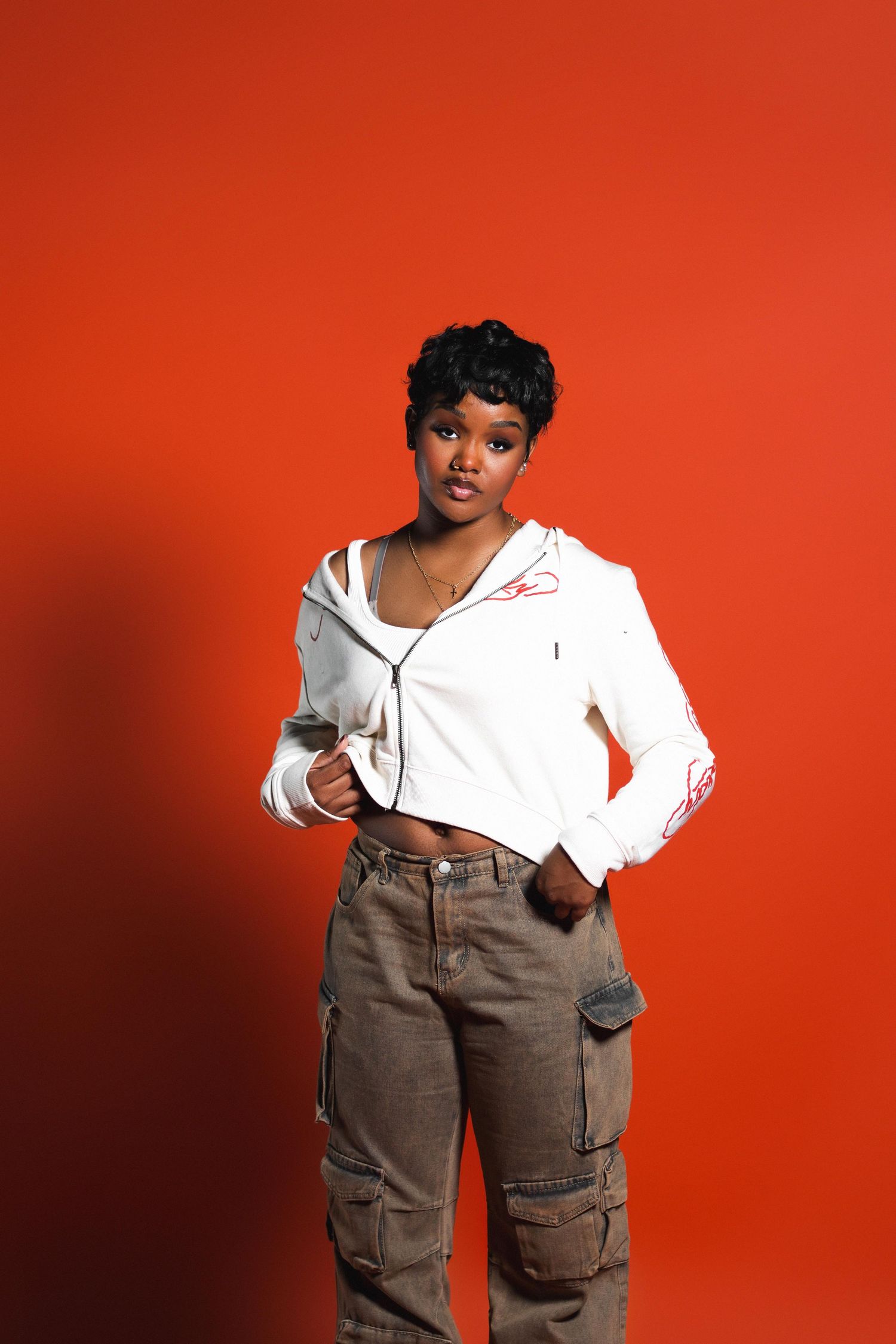 Black woman with short hair wearing white cropped hoodie and brown cargo pants on orange background.