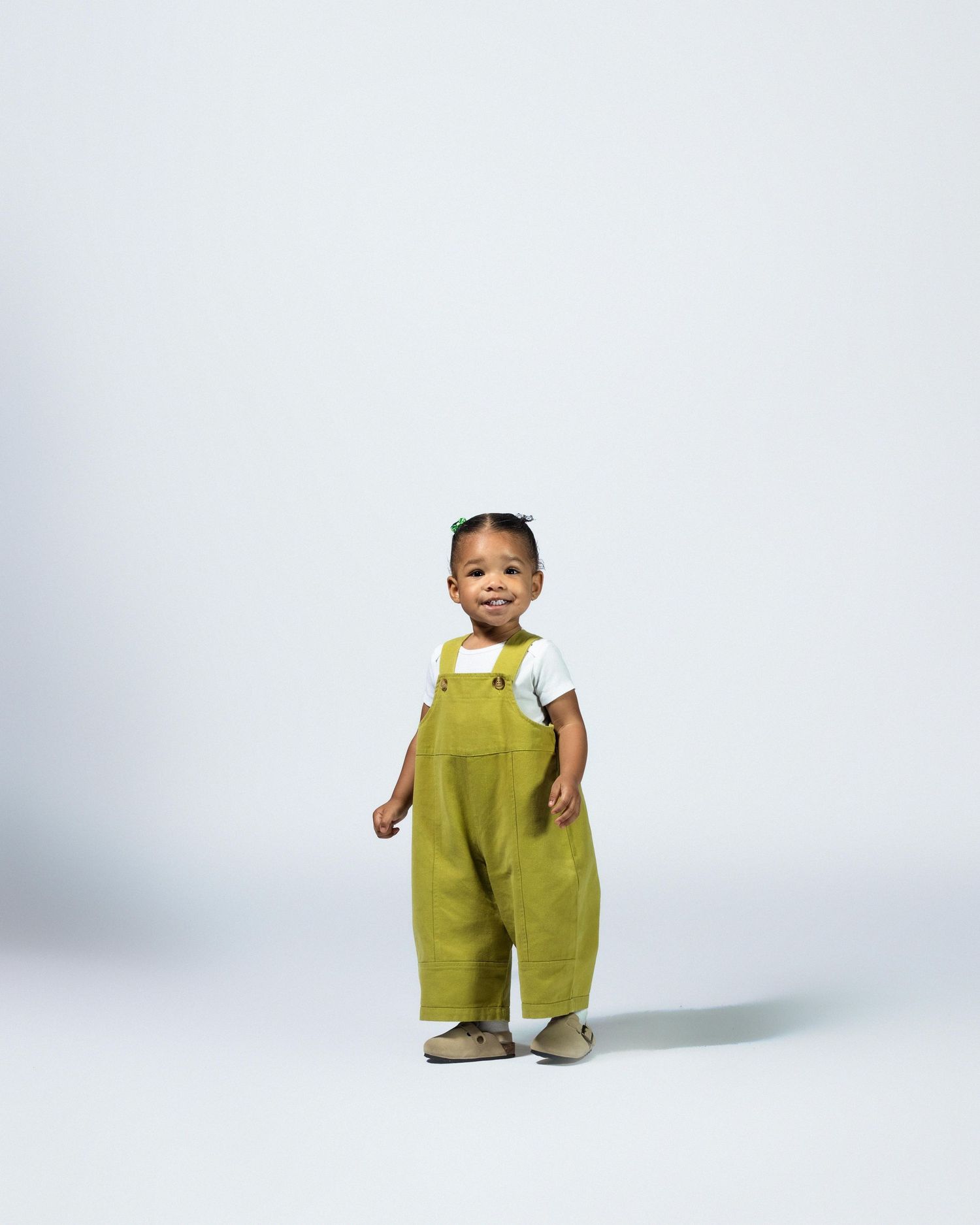 Adorable toddler in green overalls and white shirt standing on light gray background.