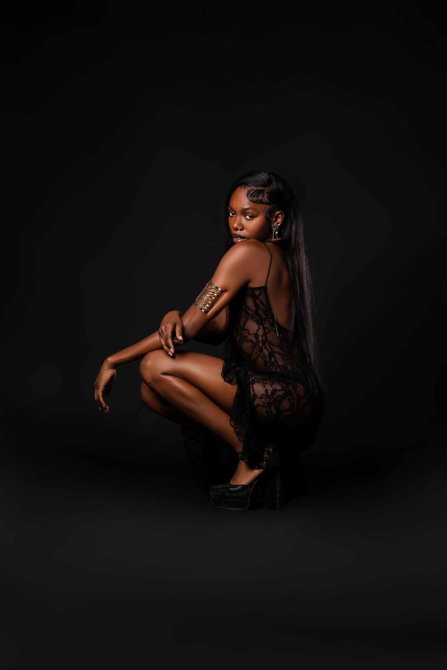 Black woman in black lace dress crouching gracefully against a dark studio background.