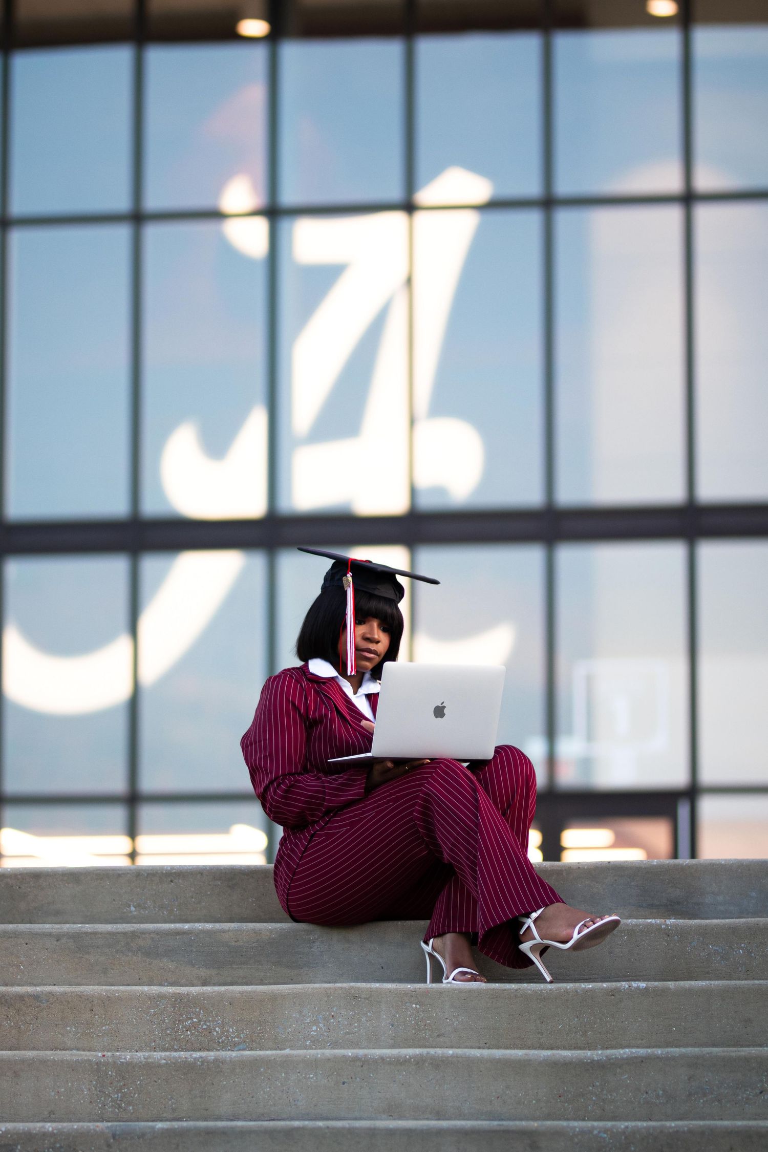 Graduate in cap and maroon pinstripe suit sits on steps with MacBook laptop in front of large university building.