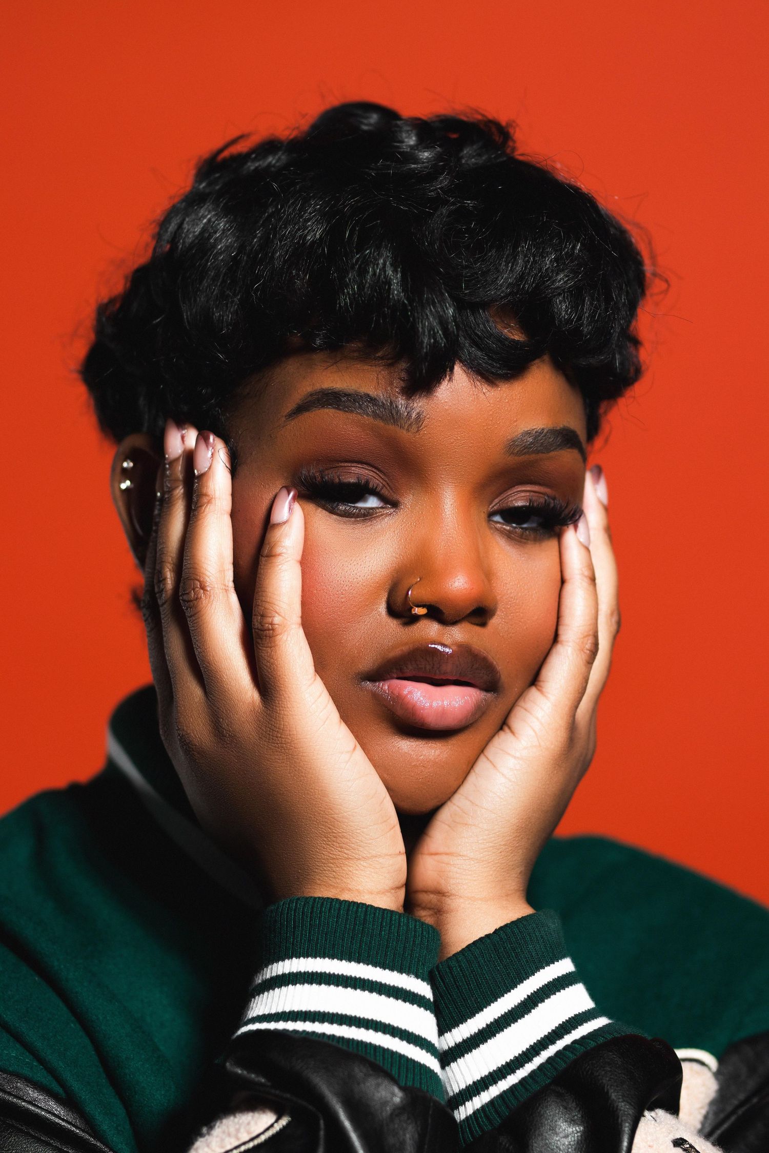 Young Black woman with short curly hair, nose ring, and green varsity jacket cradling face with both hands on red background.