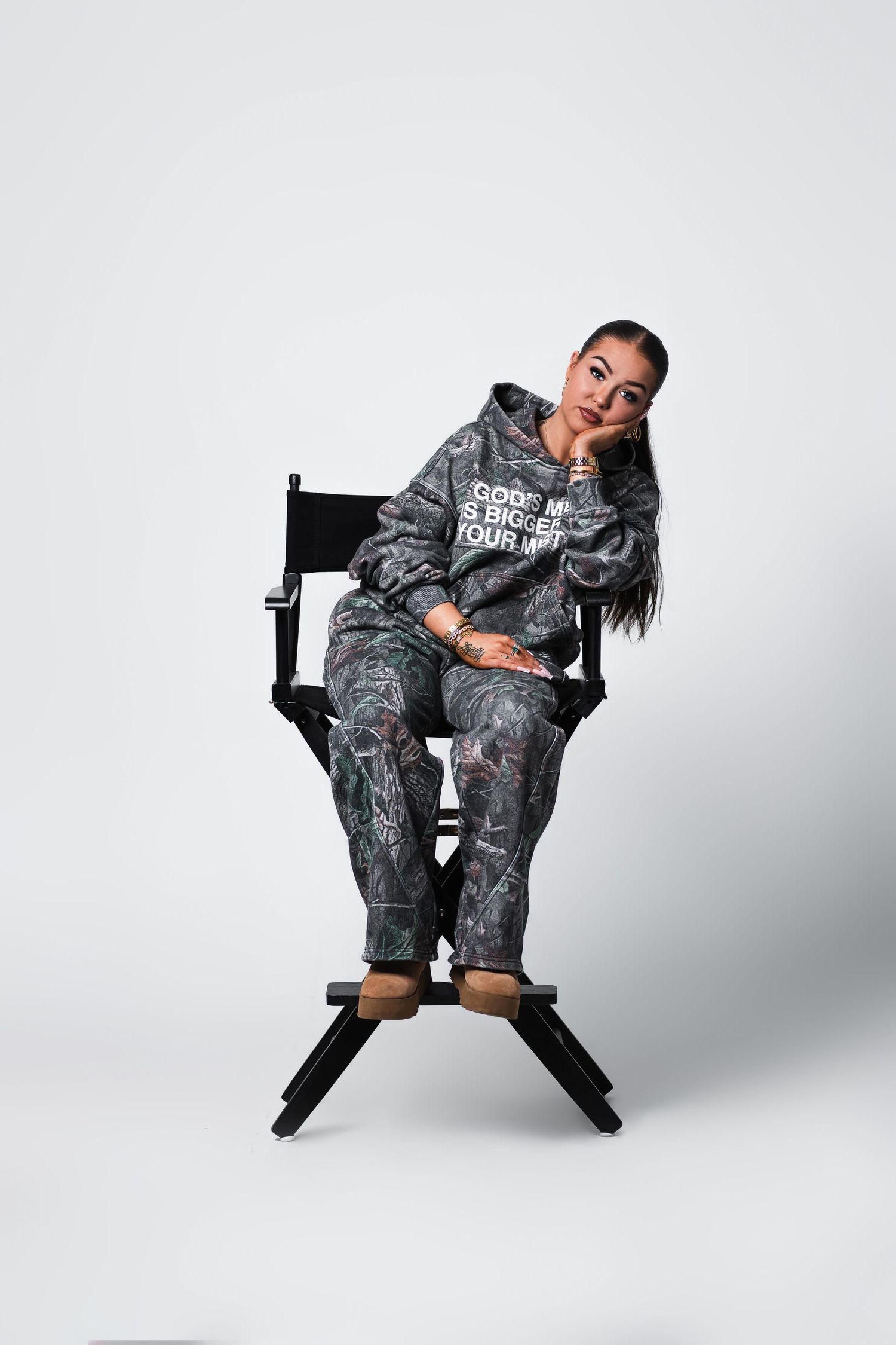 Woman in camo hoodie and pants sitting on directors chair against white background.