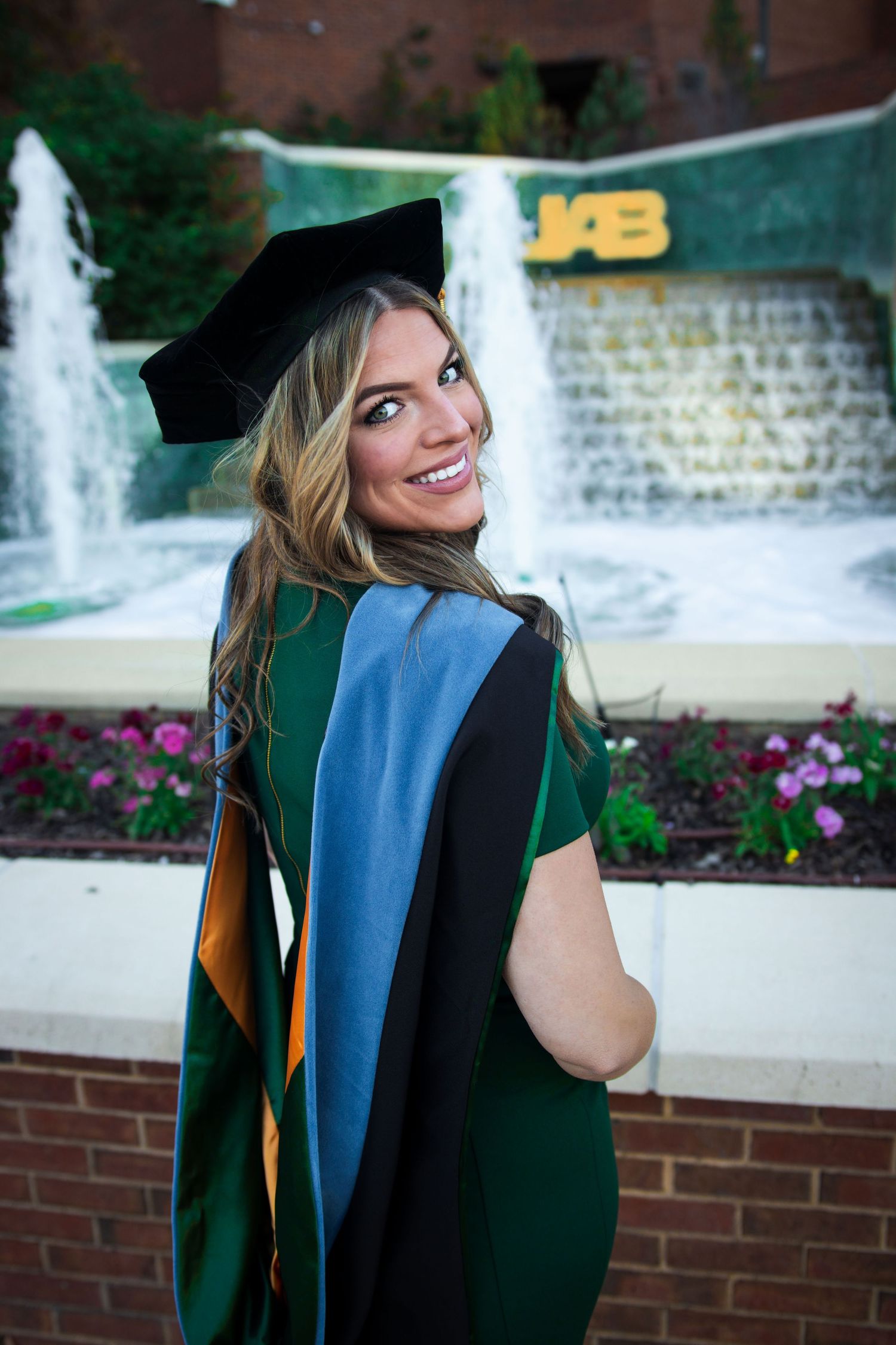 Smiling female graduate in green cap and gown poses in front of UAB fountain.