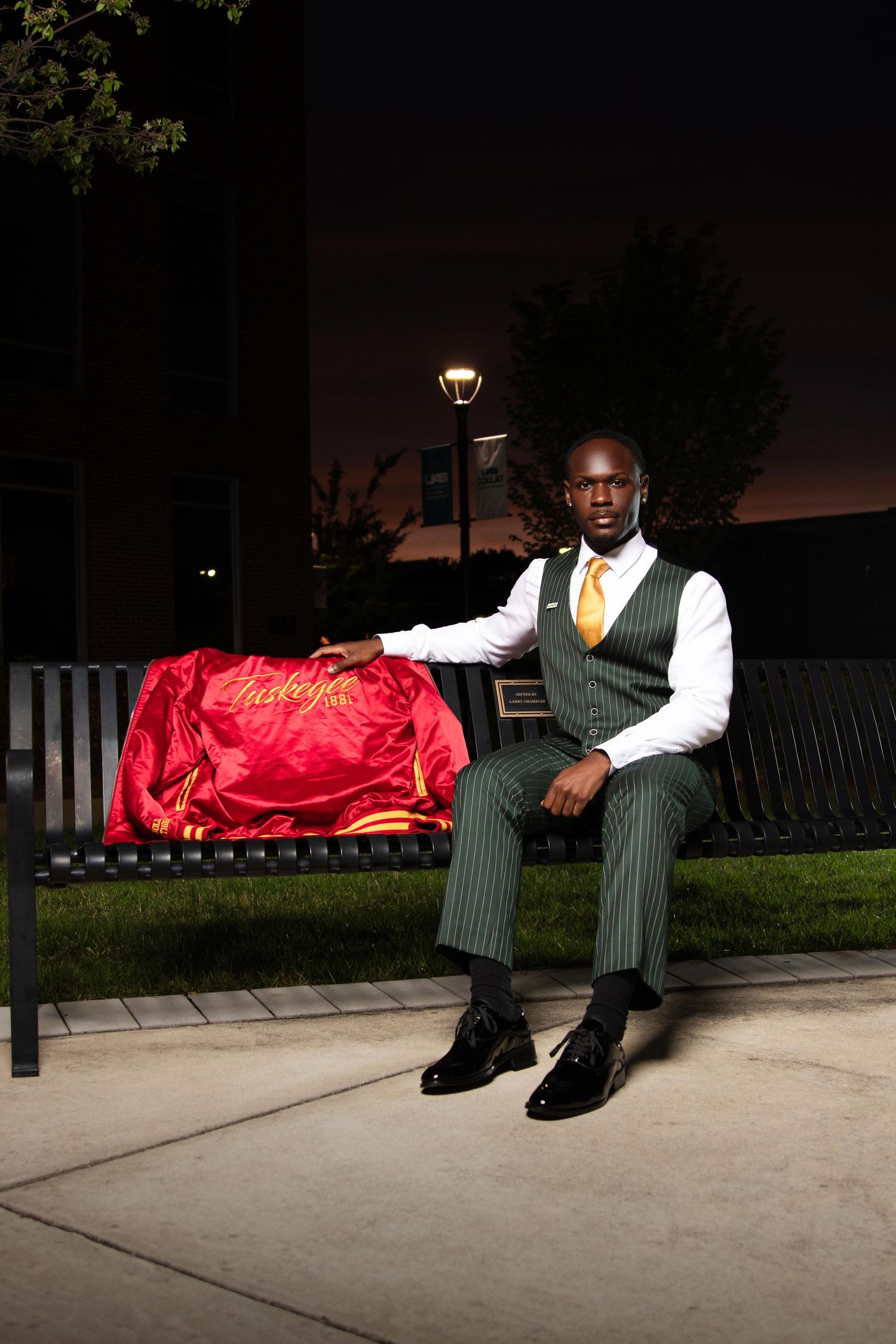 Man in green pinstripe suit sits on bench at night holding red satin jacket outdoors.