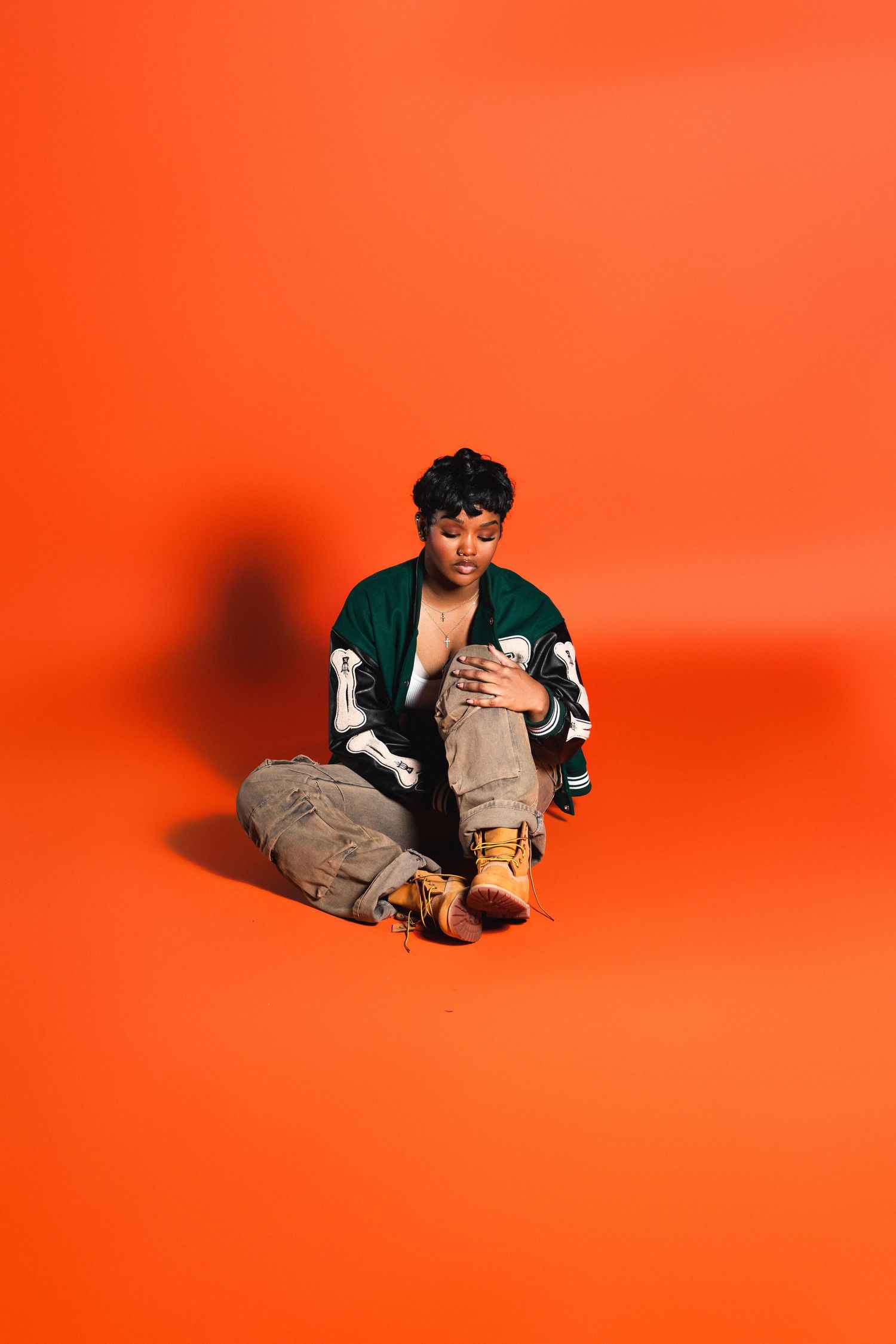 Person in green varsity jacket, cargo pants, and tan boots sitting cross-legged on orange background.
