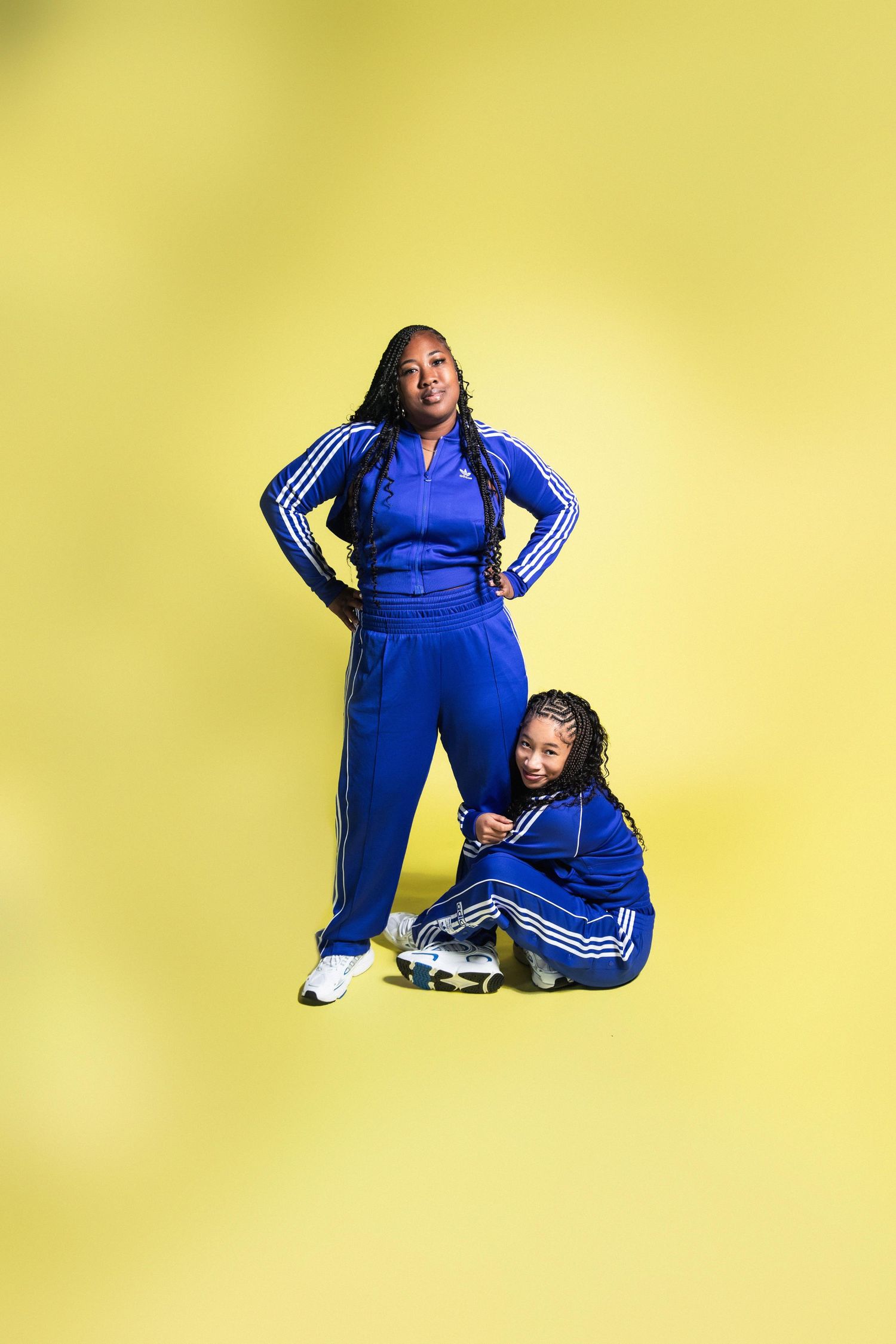 Two Black women in matching blue Adidas tracksuits pose together against a bright yellow background.