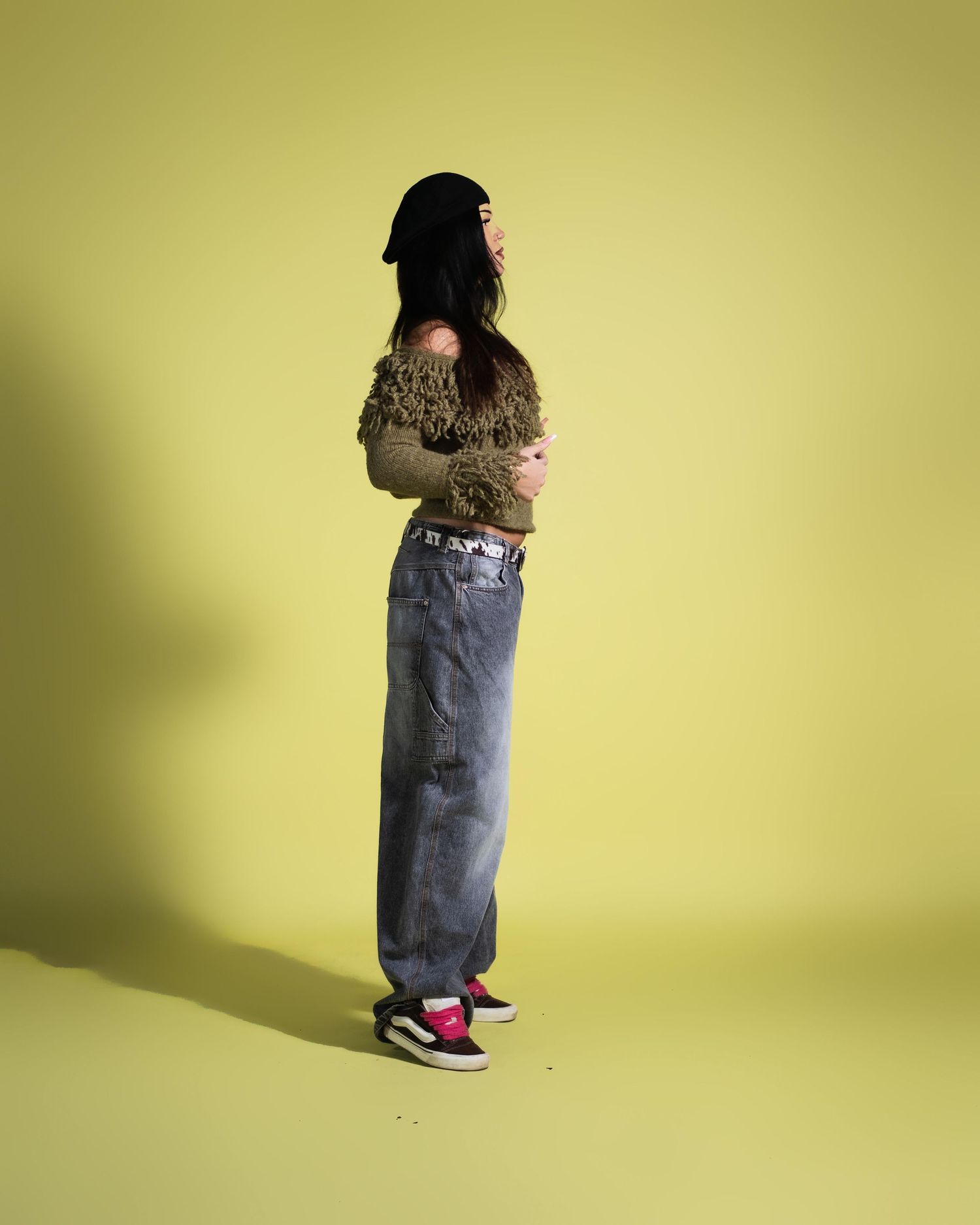 Woman in olive fringe sweater, grey baggy jeans, and red sneakers posing sideways on yellow background.