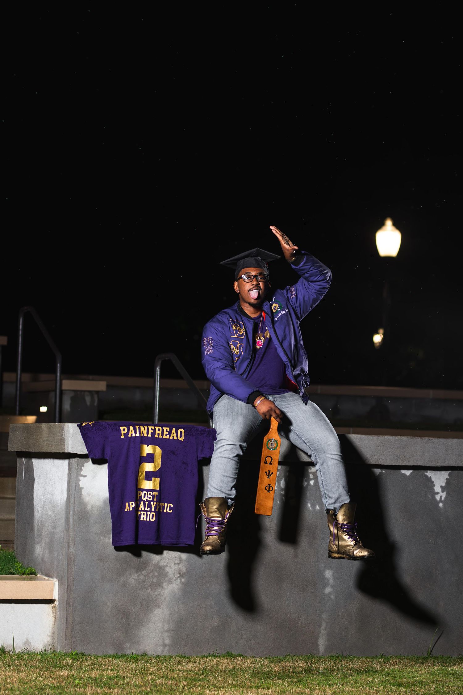 A graduate in cap and purple jacket sits on a wall at night displaying a jersey and wooden paddle.