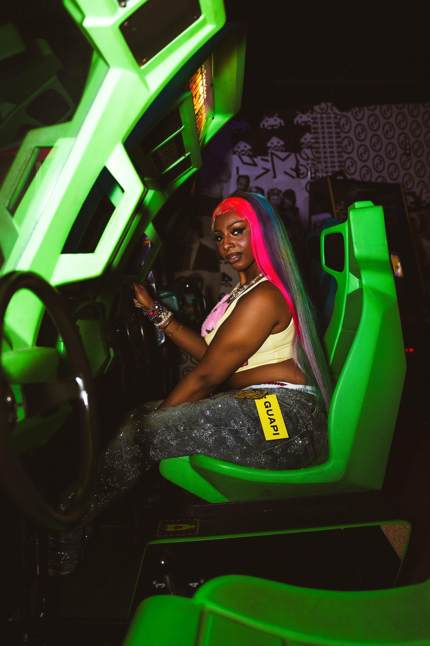 Woman with pink and blue hair sits in green neon-lit arcade racing game seat.