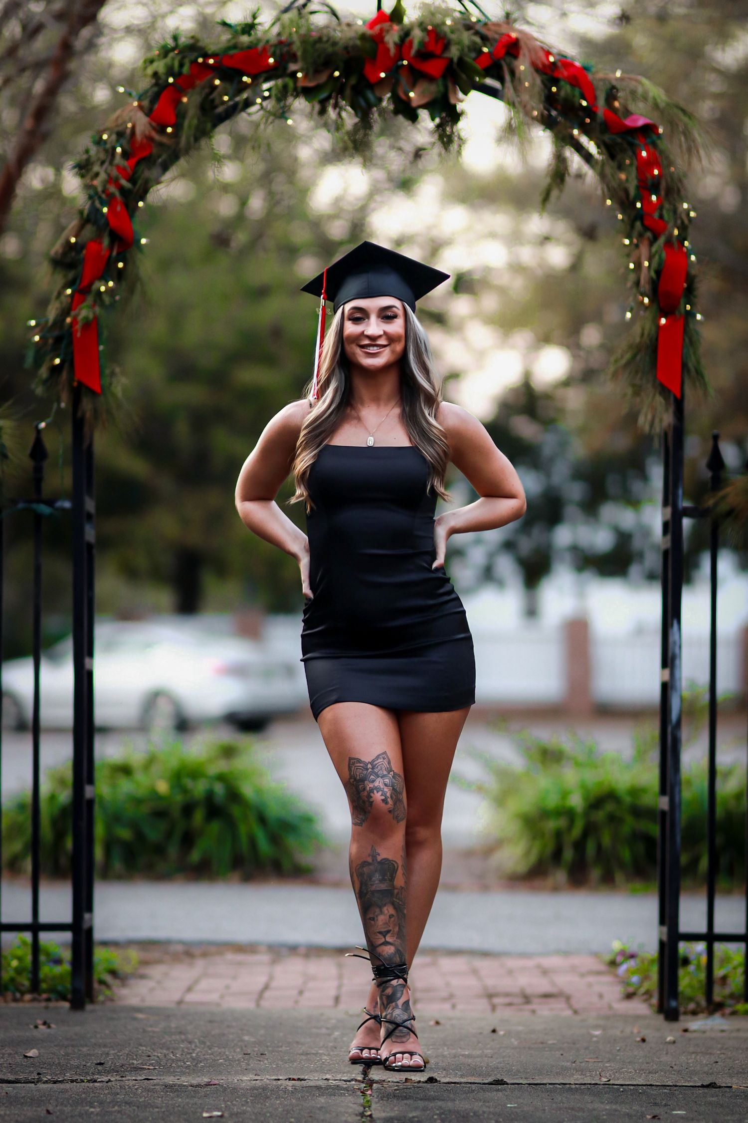 Young woman in black mini dress and graduation cap poses under festive holiday arch outdoors.
