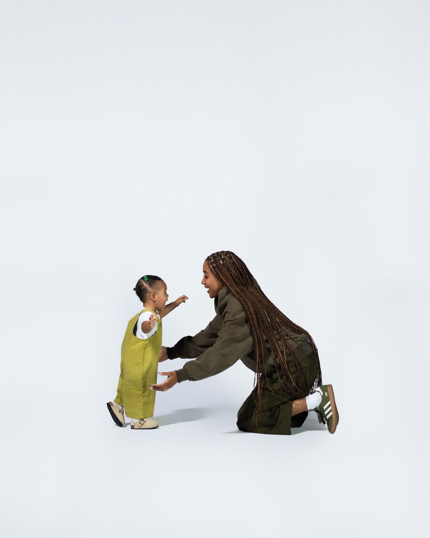 Woman with braids kneeling to interact with toddler on white background.