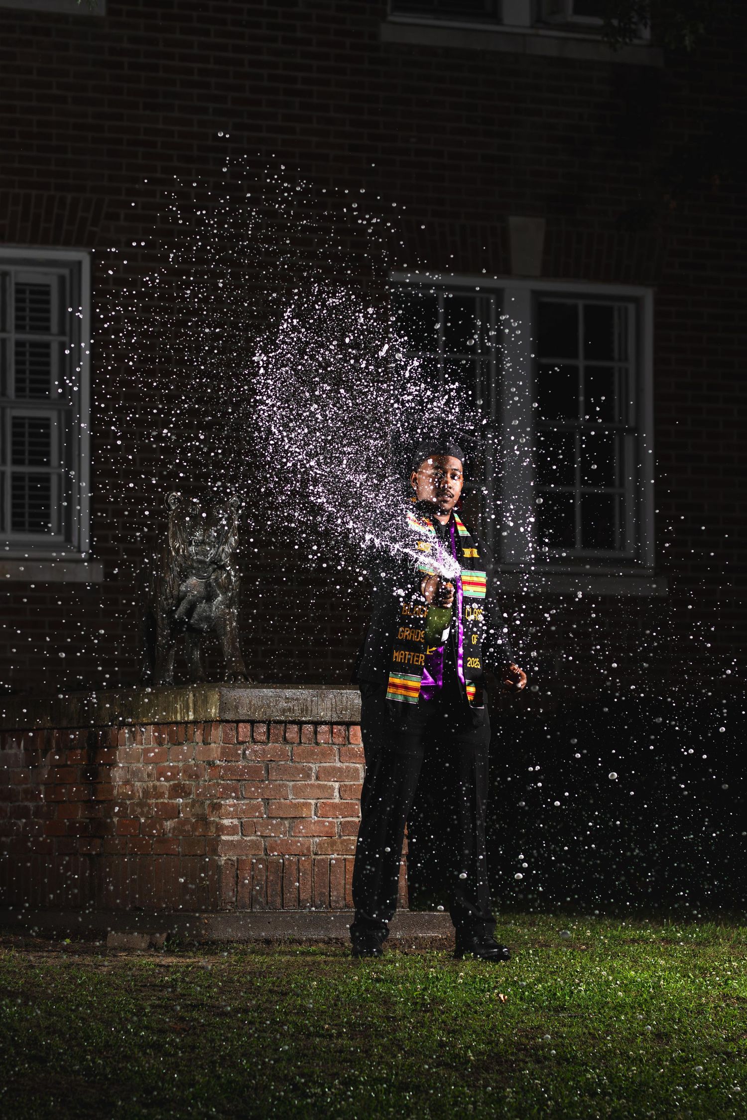 Graduate in academic regalia spraying champagne at night outside a brick building with dramatic effect.