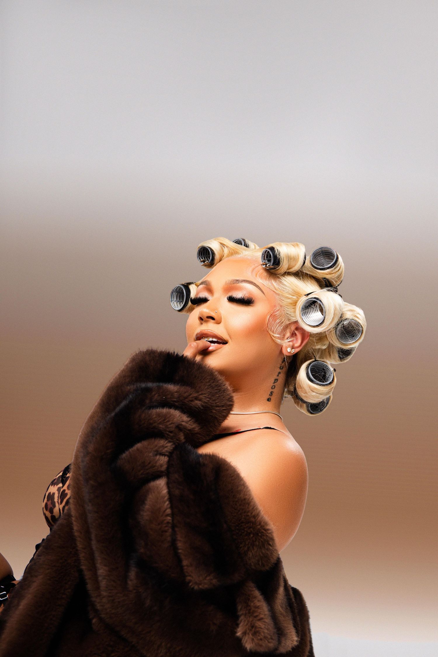 Woman with blonde hair in rollers wearing a brown fur coat and leopard print top poses against a neutral background.