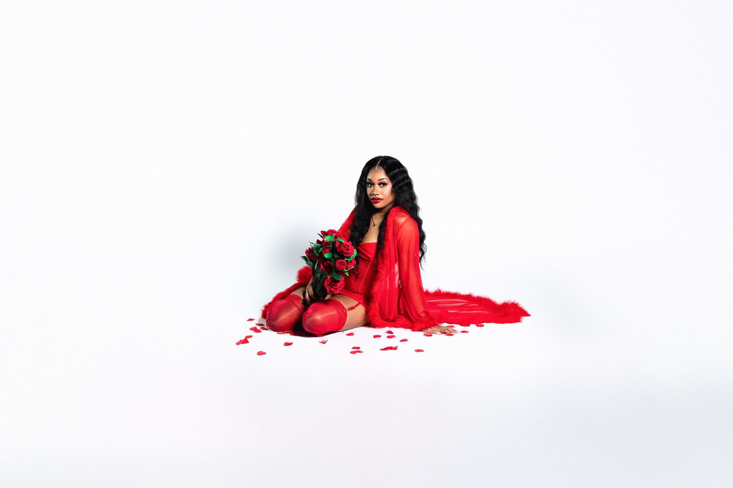 Woman in red sheer dress sitting on floor with red roses and scattered petals on white background.
