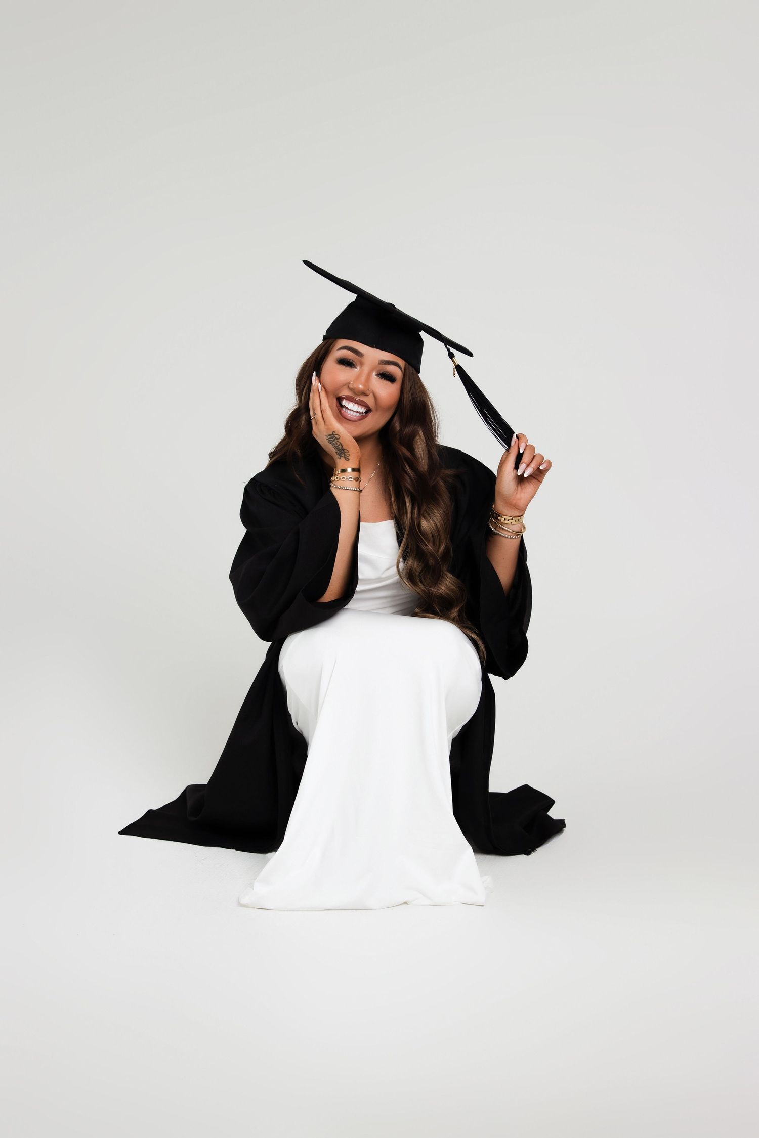 Joyful graduate in black cap and gown seated in white dress smiling on a light gray background.