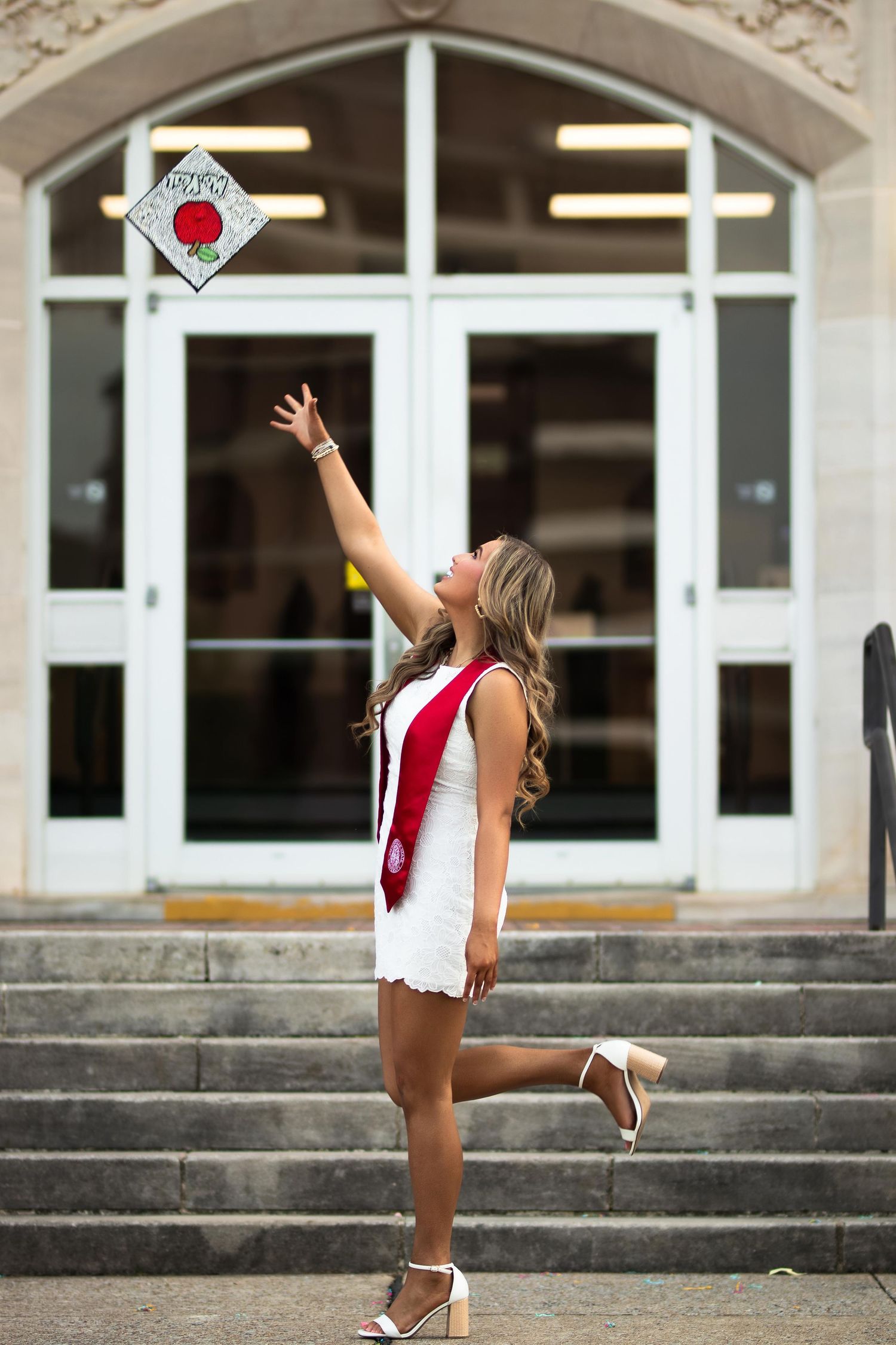 A graduating woman in a white dress and red sash tosses her decorated cap in front of building doors.
