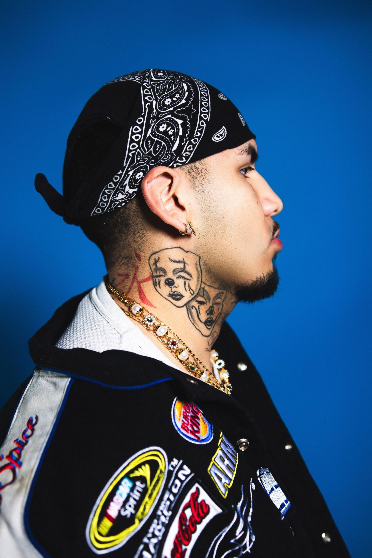 A profile view of a tattooed man with a bandana and neck tattoos wearing a NASCAR-style jacket.