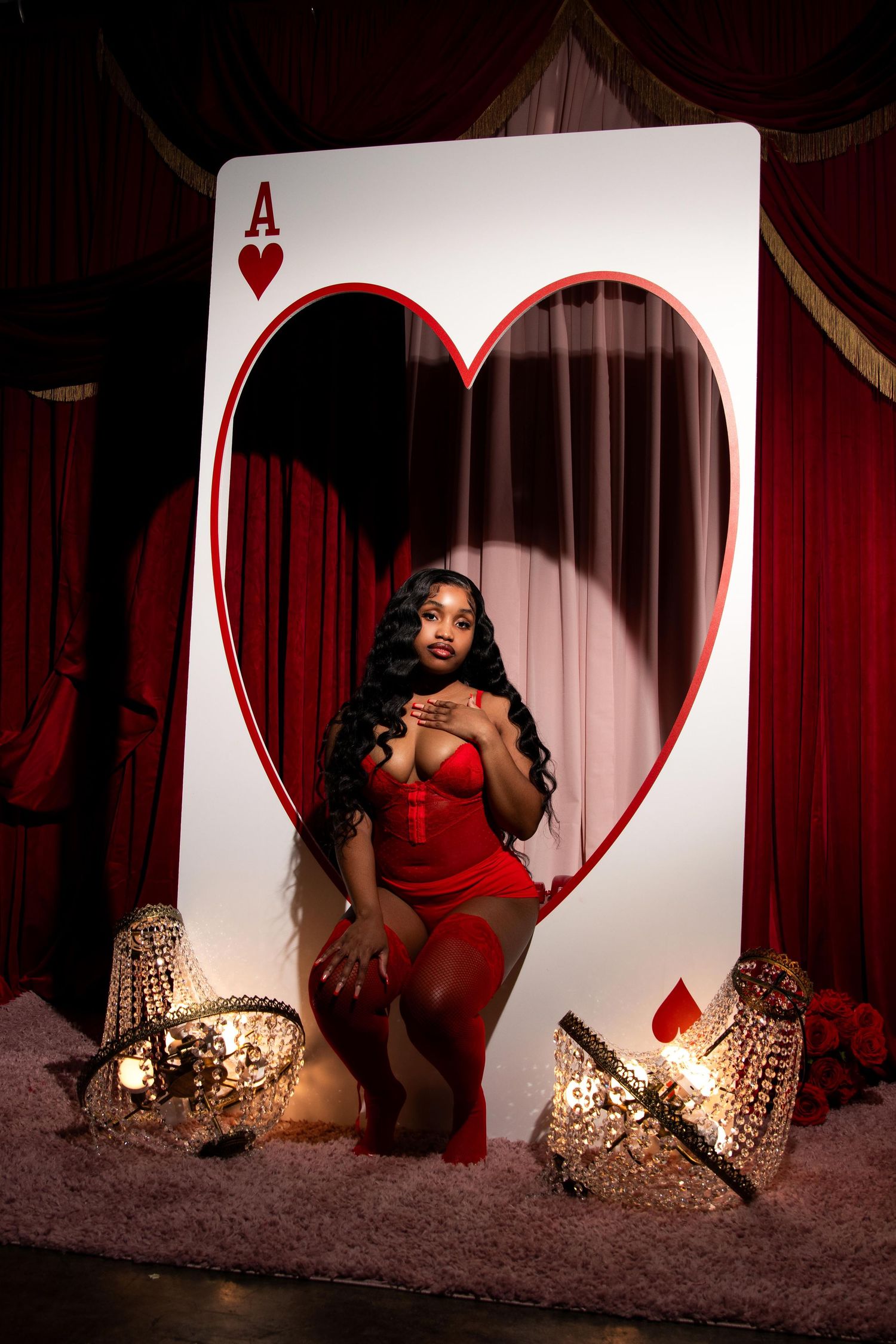 Woman in red lingerie poses in front of oversized Ace of Hearts card with crystal decorations and red curtain backdrop.