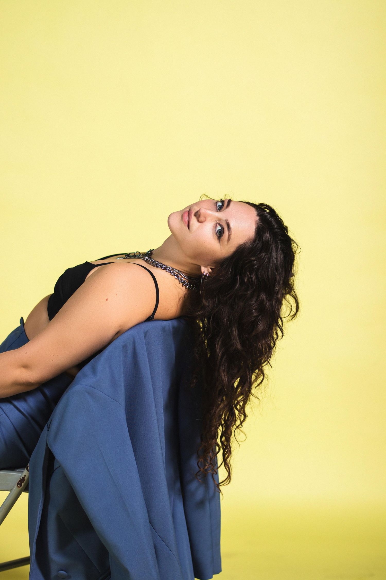 Brunette woman with curly hair leaning back on chair in blue pants and black top, yellow background.