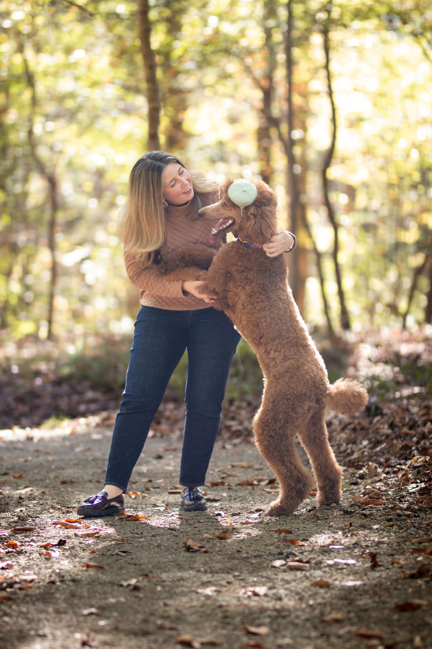 Woman in brown sweater playing with an apricot standard poodle jumping up on a forest trail in autumn.